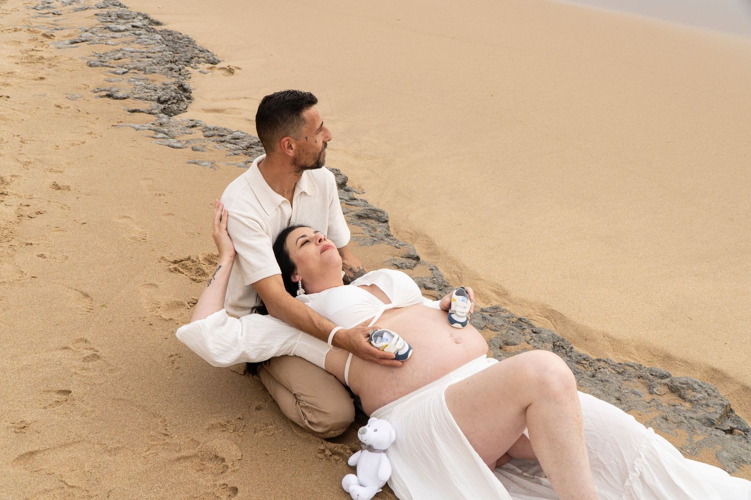 Pregnant couple relaxing on sandy beach, woman lying down with baby shoes on belly, man sitting beside her.