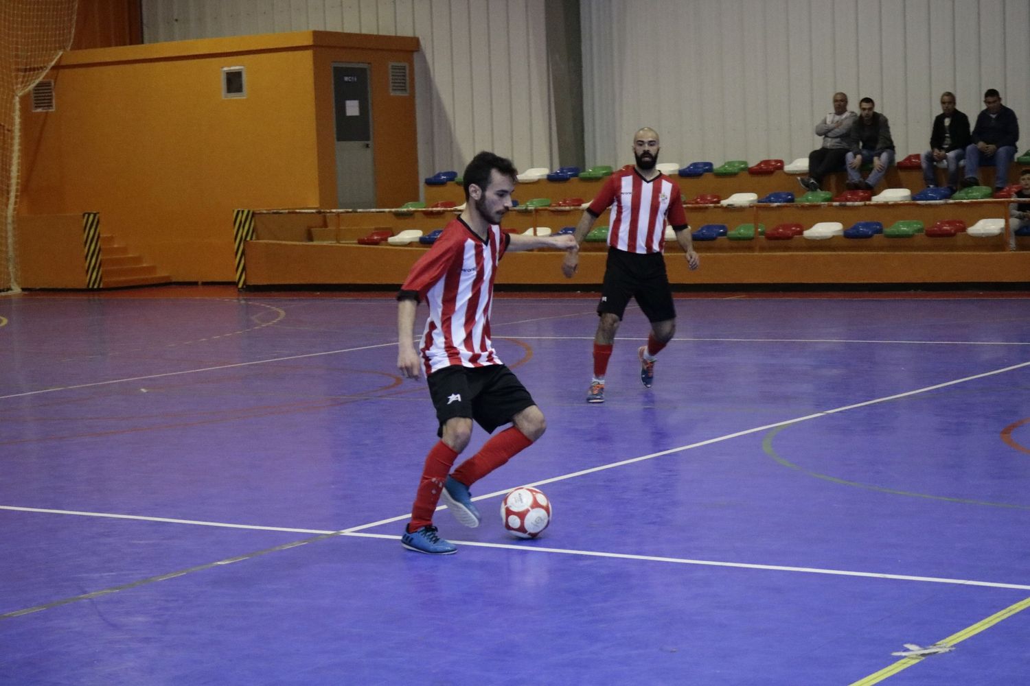 Soccer player in red and white striped jersey dribbles ball on indoor purple court during futsal match.