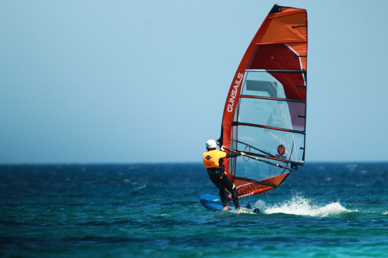Windsurfer with red sail gliding across turquoise waters on a bright day.