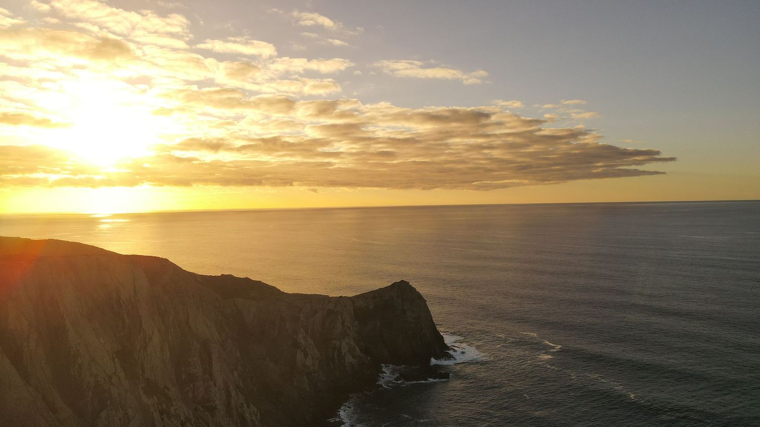 Dramatic coastal cliff silhouetted against golden sunset sky over calm ocean waters.