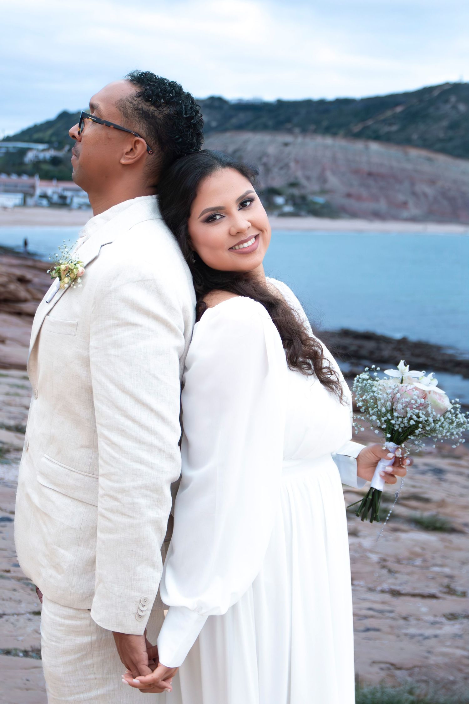 A couple in matching white outfits share romantic moments on a scenic beach overlooking the water and cliffs.