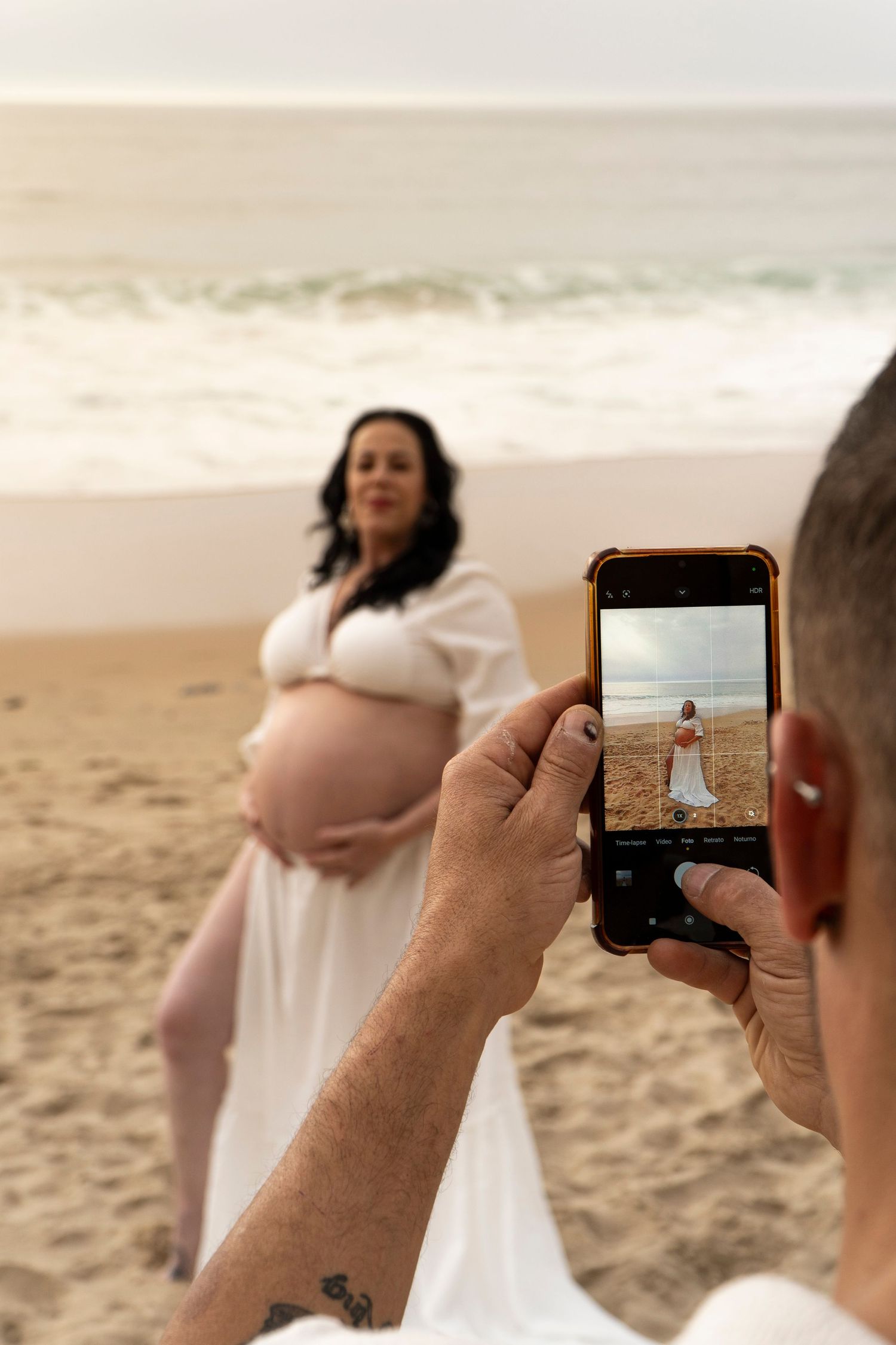 Man photographs pregnant woman in white dress on beach, capturing maternity moment with ocean waves in background.