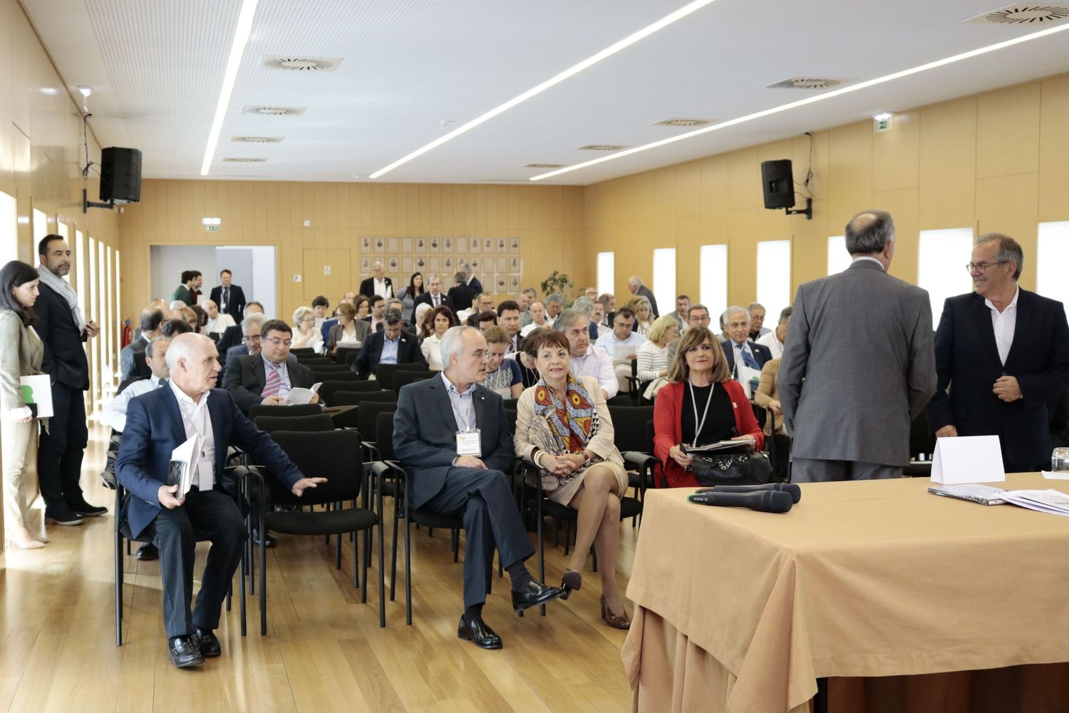 People gathered in a brightly lit conference room with rows of chairs and tables during a formal meeting or event.