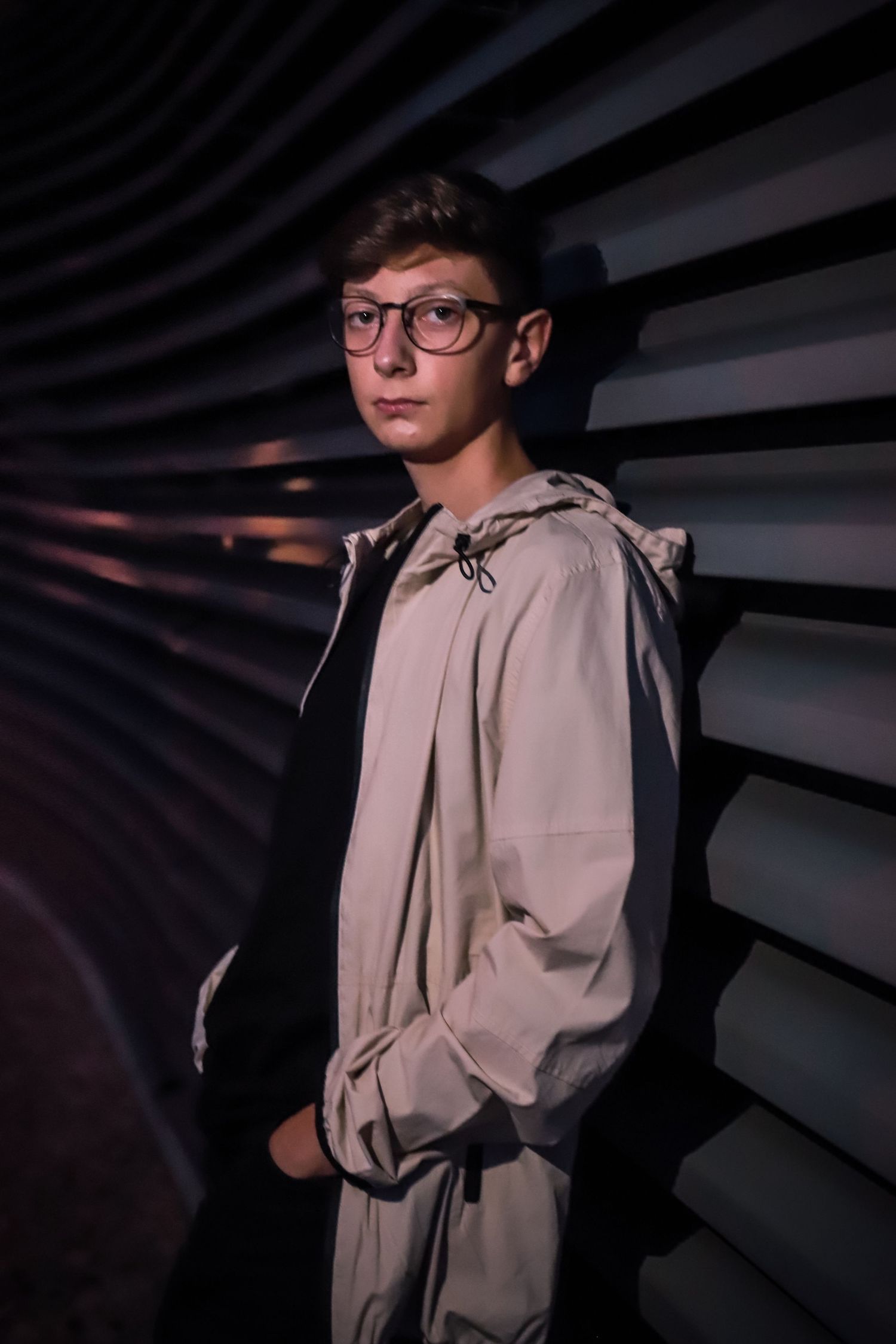 Person in a light hoodie and glasses posing against a striped wall with dramatic lighting.