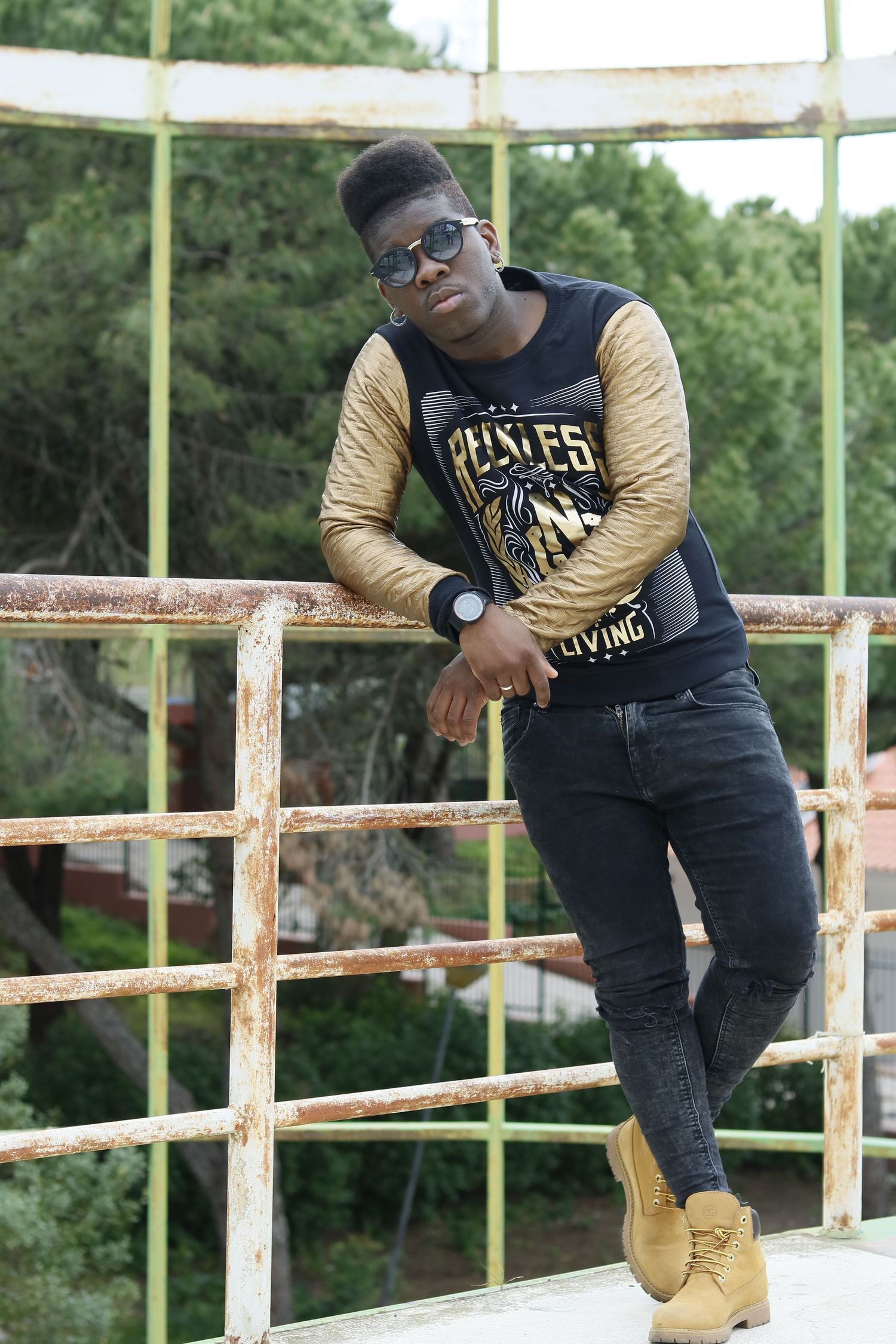 A person in casual streetwear poses on a railing outdoors wearing a black and gold varsity shirt, jeans and tan boots.