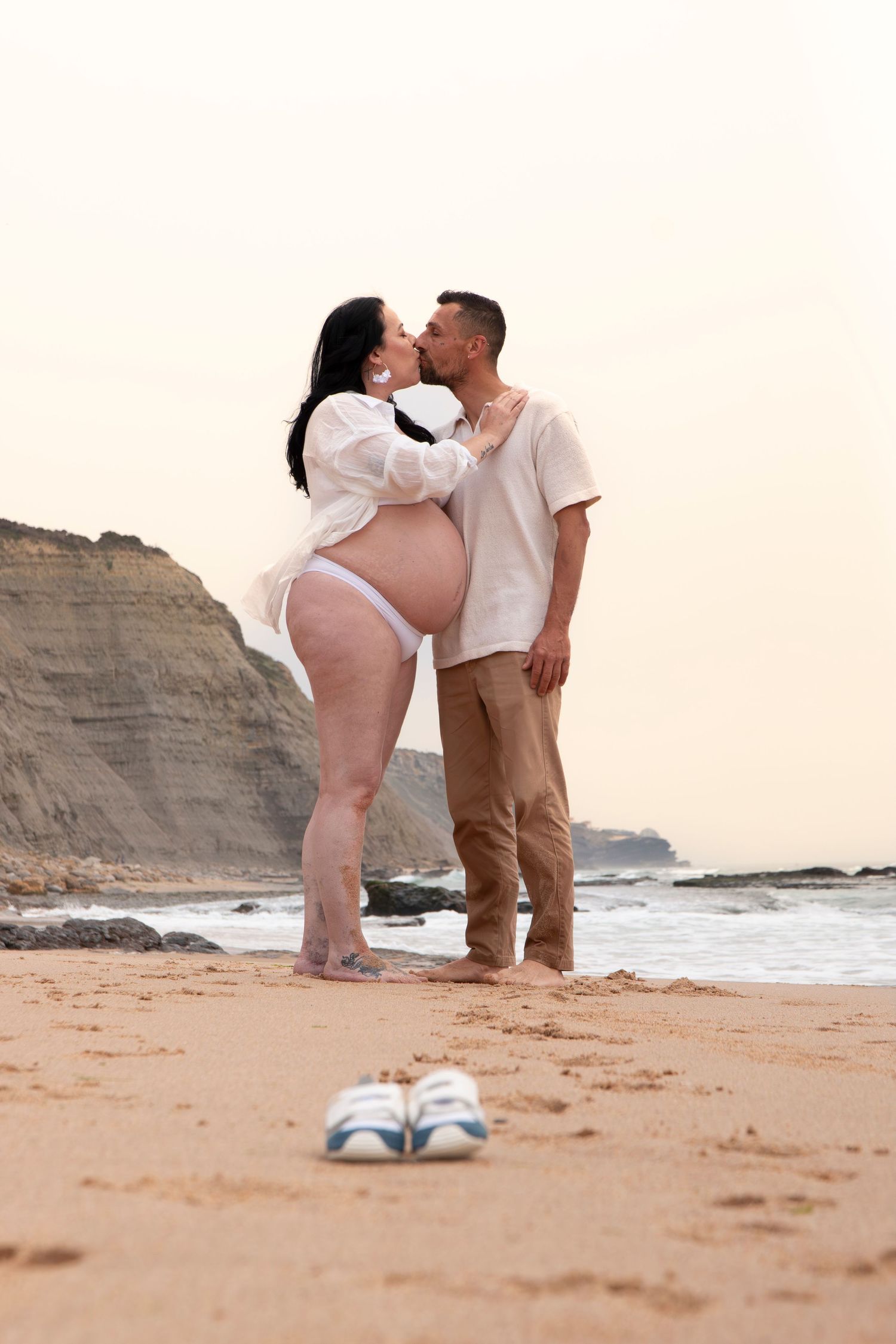 Pregnant couple sharing a kiss on a sandy beach at sunset, with baby shoes in the foreground.