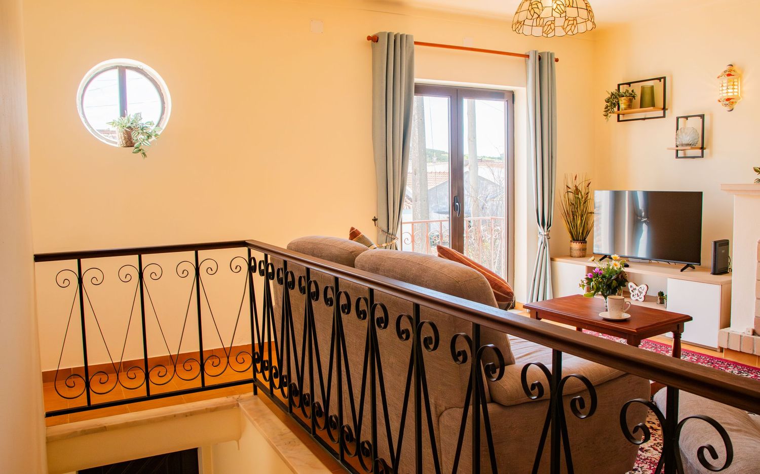A cozy split-level living room with wrought iron railings, peach walls, gray curtains, and a round window.