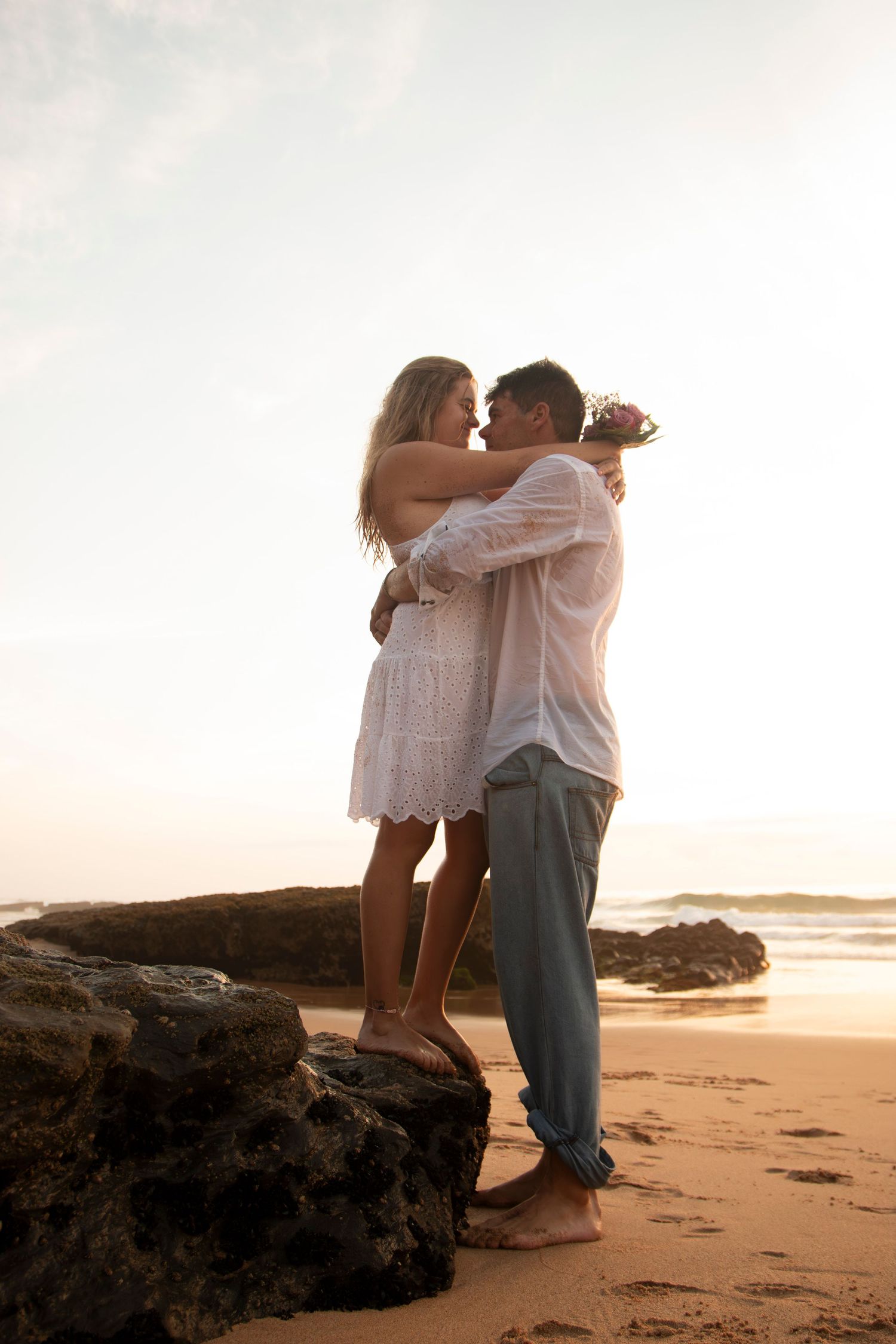 A couple shares an intimate embrace on a rocky beach at sunset creating a romantic silhouette against the ocean backdrop.
