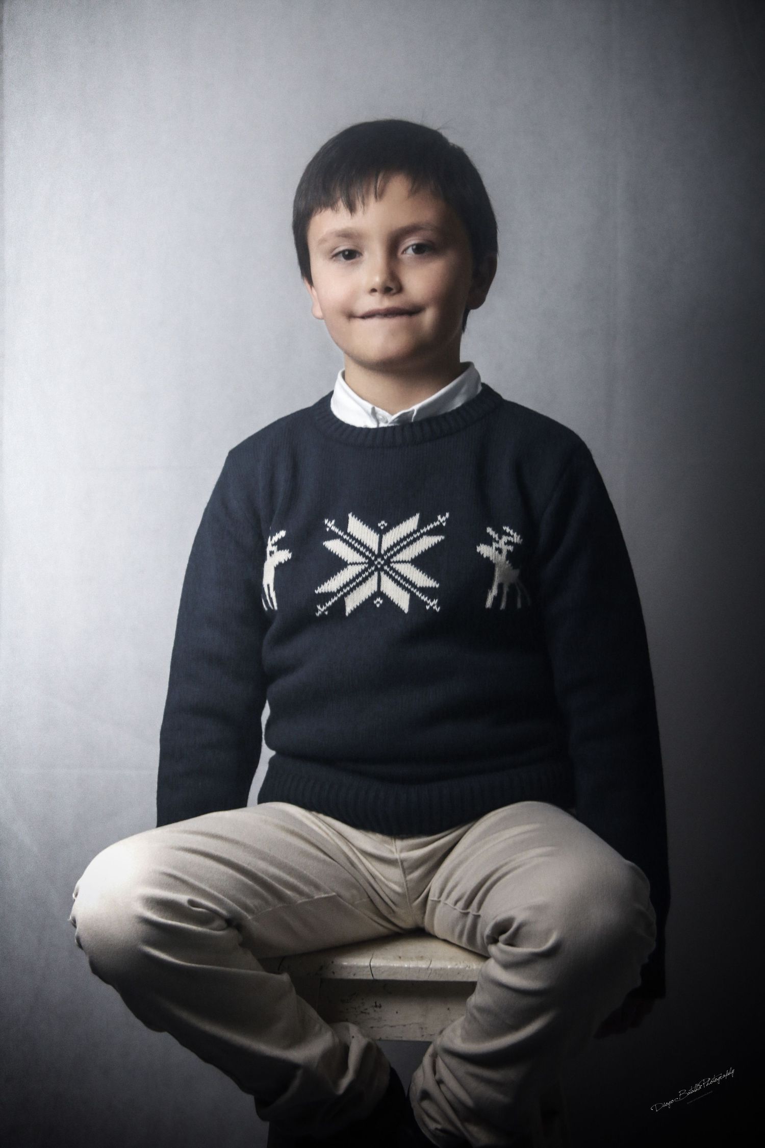 Portrait of child wearing navy winter sweater sitting cross-legged against grey backdrop.