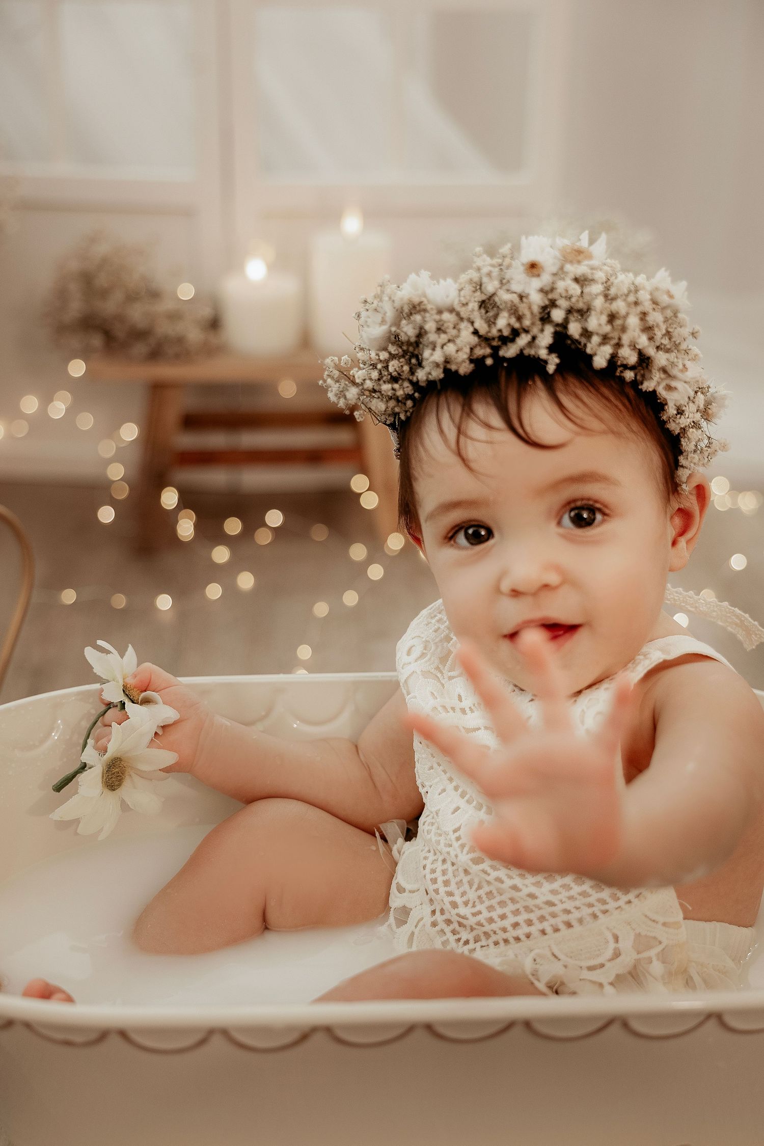 A baby in a white flower crown and pearls lies on a cream-colored blanket with twinkling fairy lights in the background.