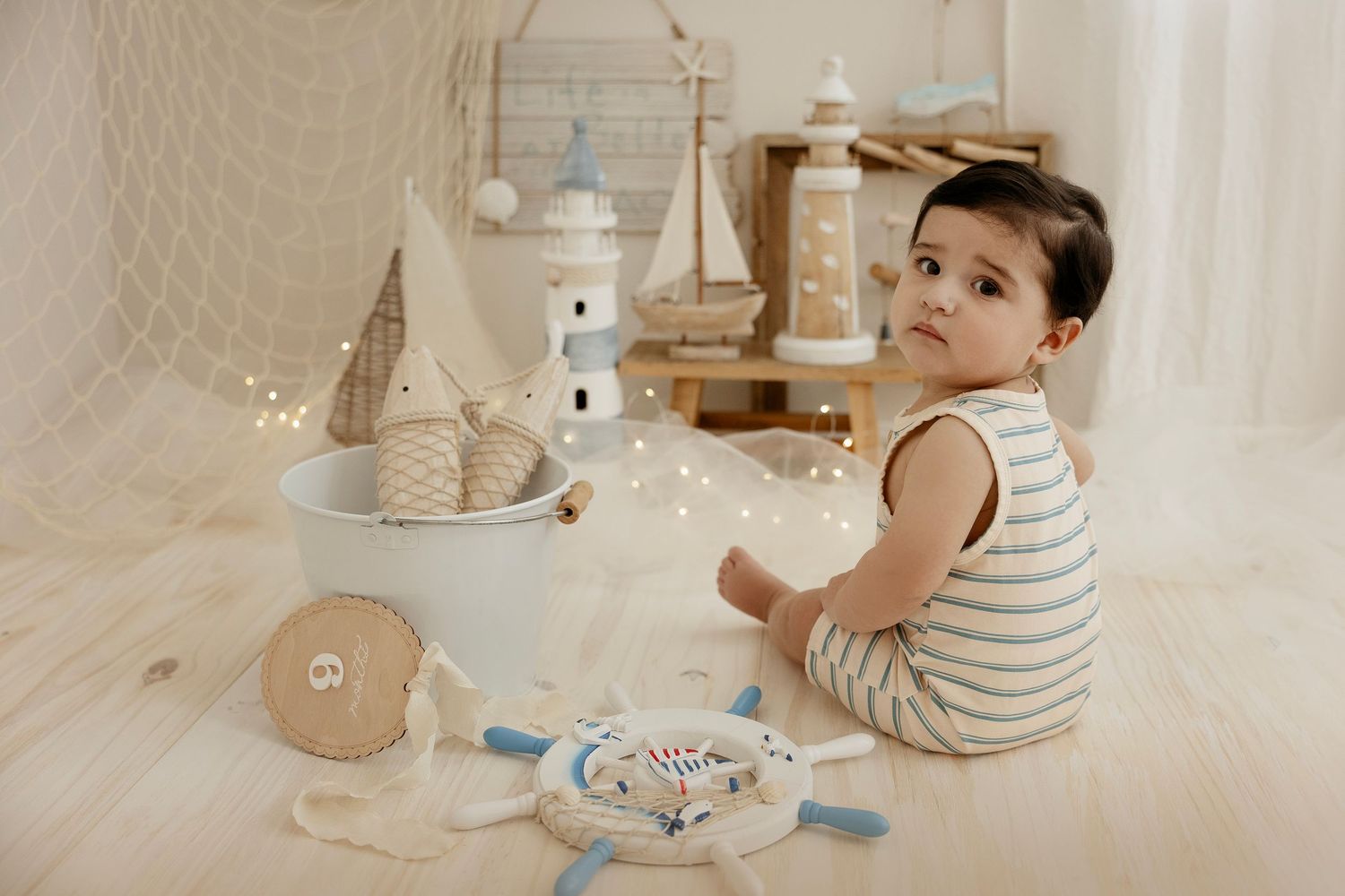 Baby in striped outfit sitting on floor with nautical toys, lighthouse decor, and fairy lights in soft white studio setting.