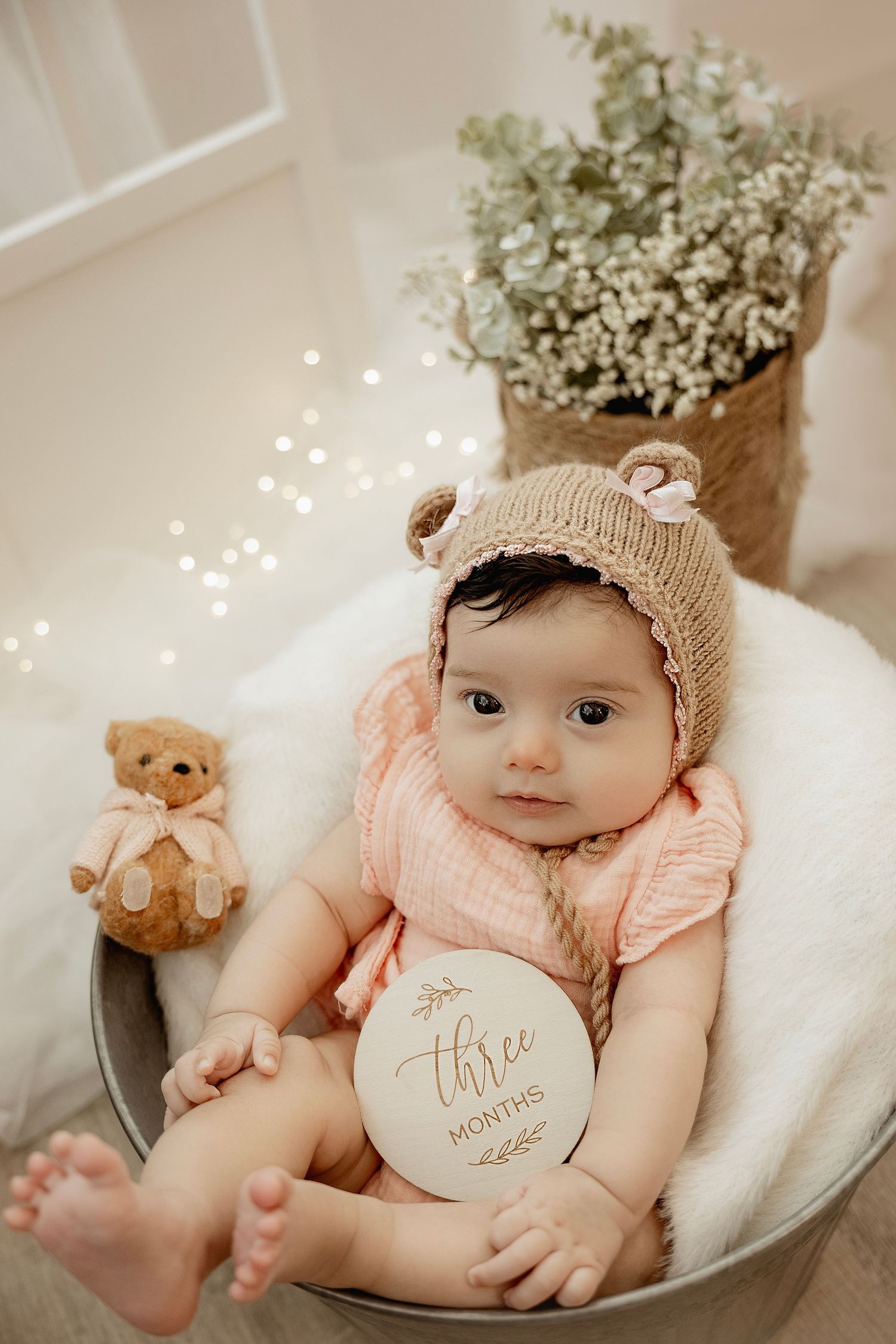 Adorable 3-month-old baby in bear bonnet and pink outfit sits in metal bucket with teddy bear and floral backdrop.