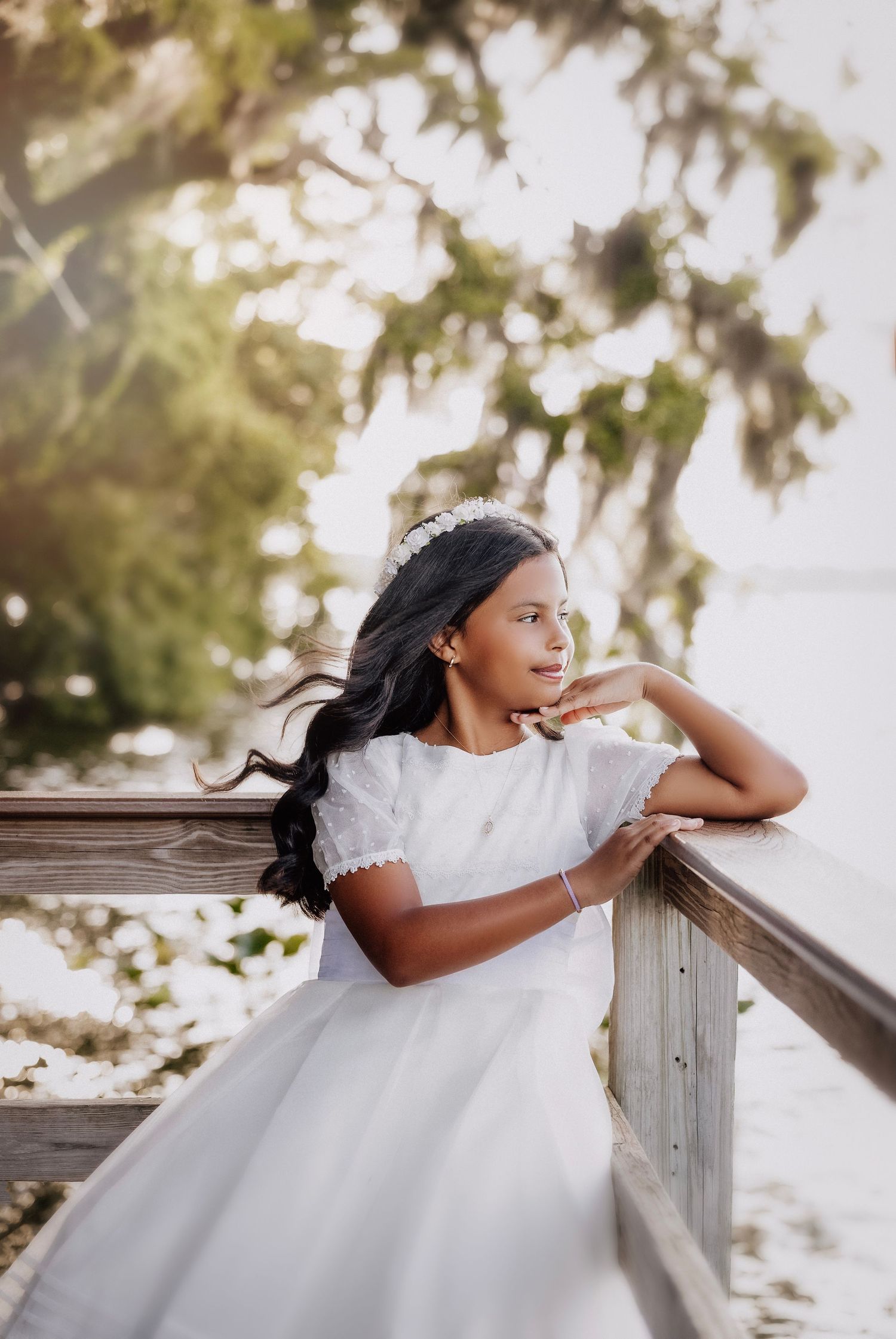 Young girl in white dress leaning on wooden railing outdoors, wearing floral headband with trees in background.