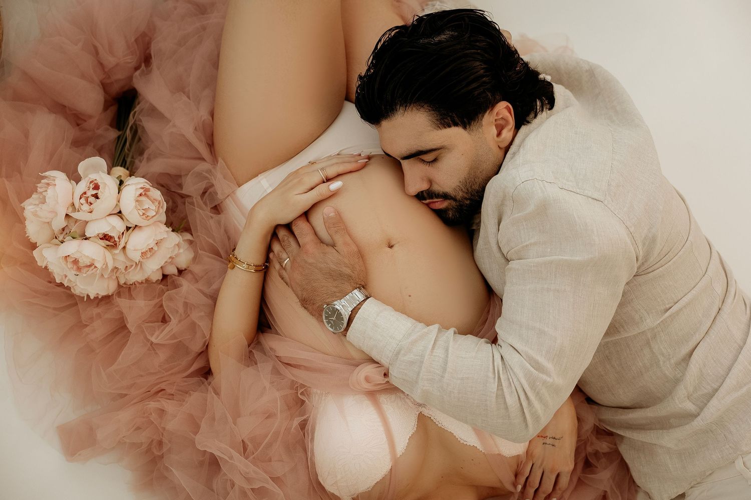 Couple sharing intimate maternity moment, man embracing pregnant woman in pink tulle skirt with roses.