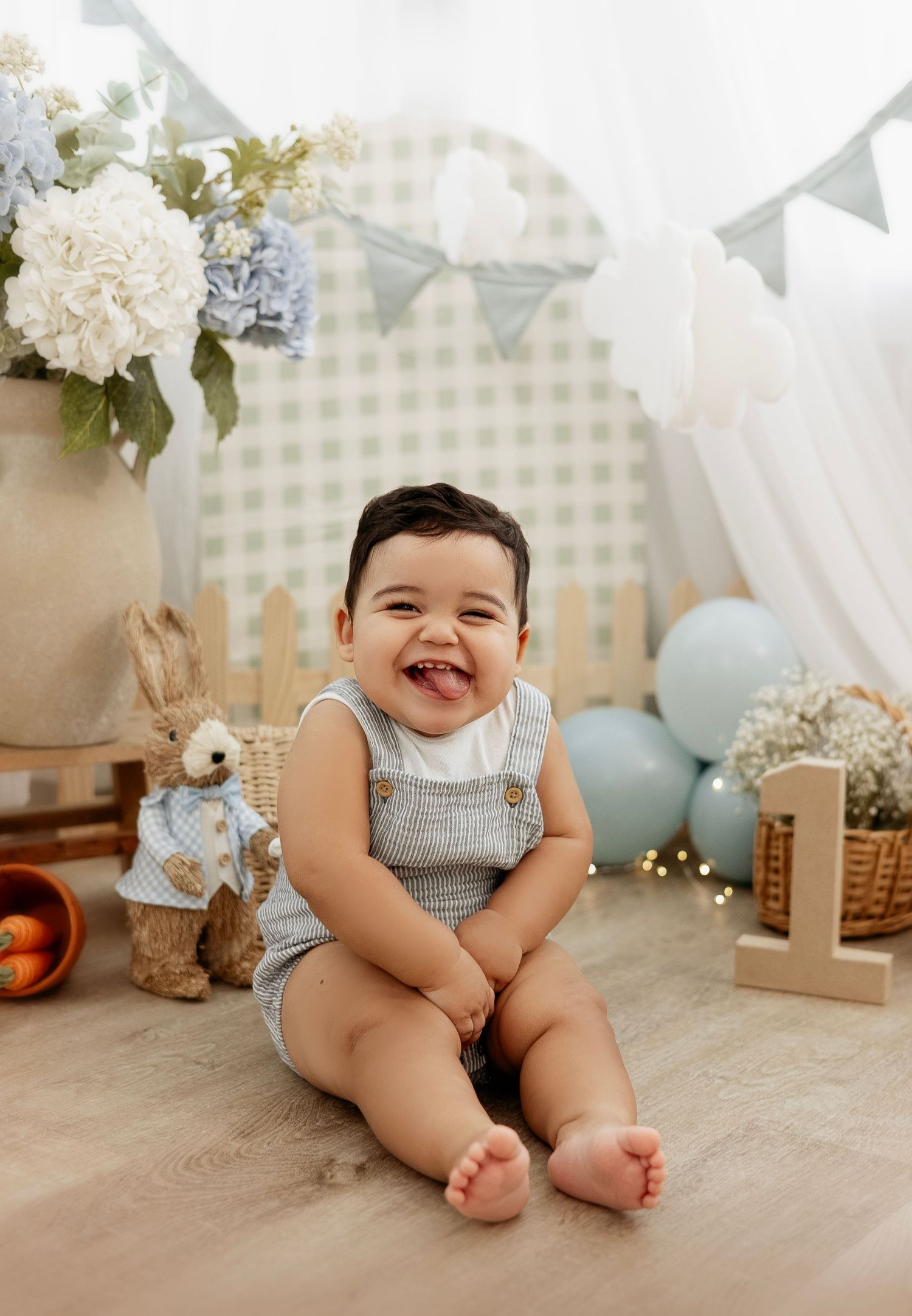 Baby wearing gray romper sits on cream colored floor with pastel decorations and floral arrangements in background.