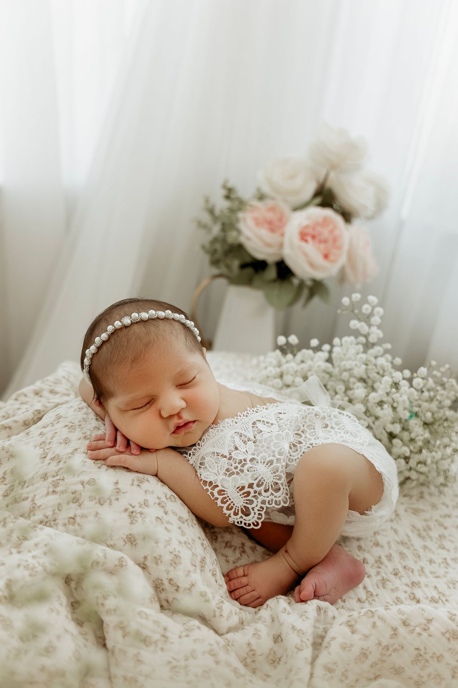 A sleeping newborn rests peacefully on white lace fabric surrounded by delicate baby's breath flowers and soft pink roses.