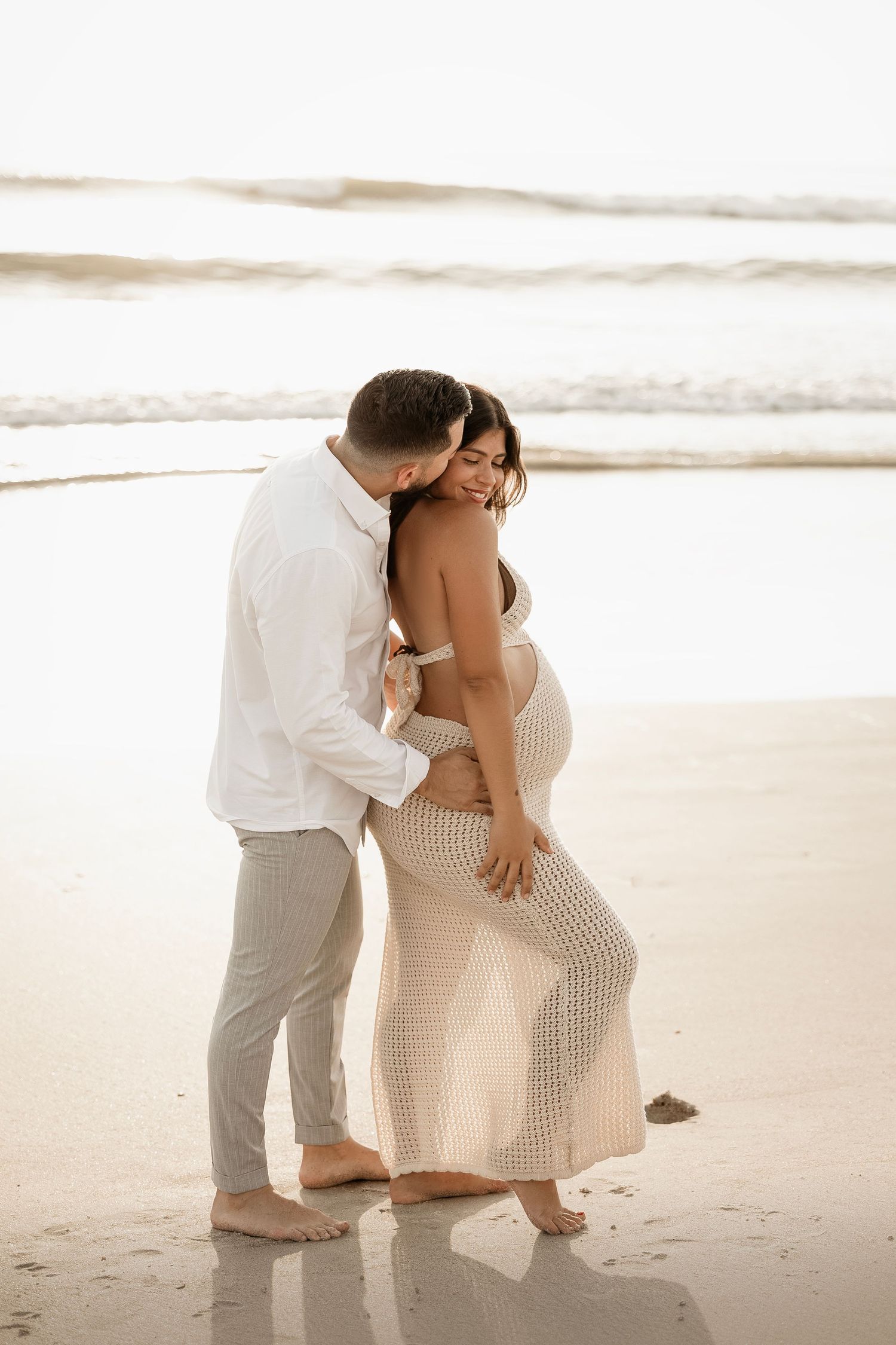 A romantic couple embraces on a serene beach at sunset wearing light colored clothing and standing barefoot in the sand.