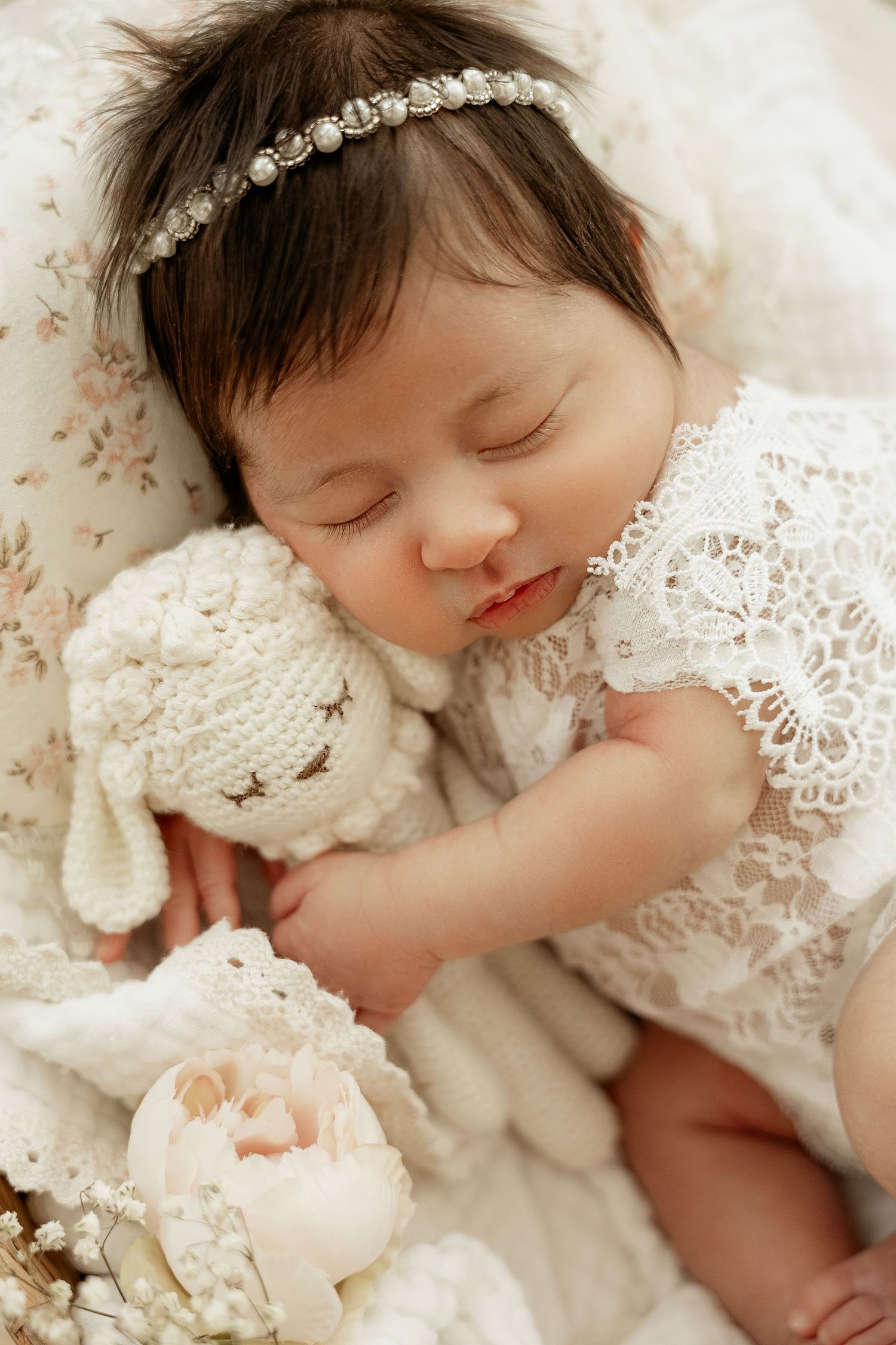 Sleeping baby wearing delicate lace dress rests peacefully on soft cream-colored bedding.