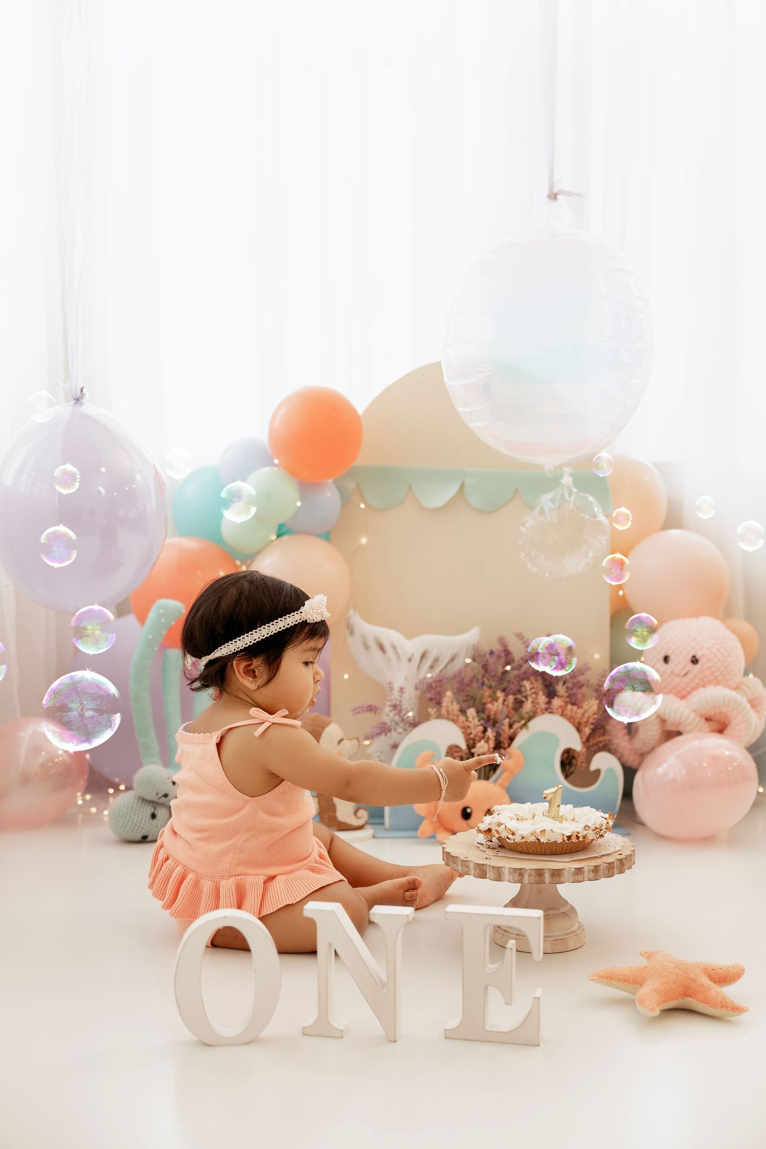 Baby girl in pink dress celebrates first birthday with cake, balloons, bubbles, and ONE sign in pastel ocean theme.