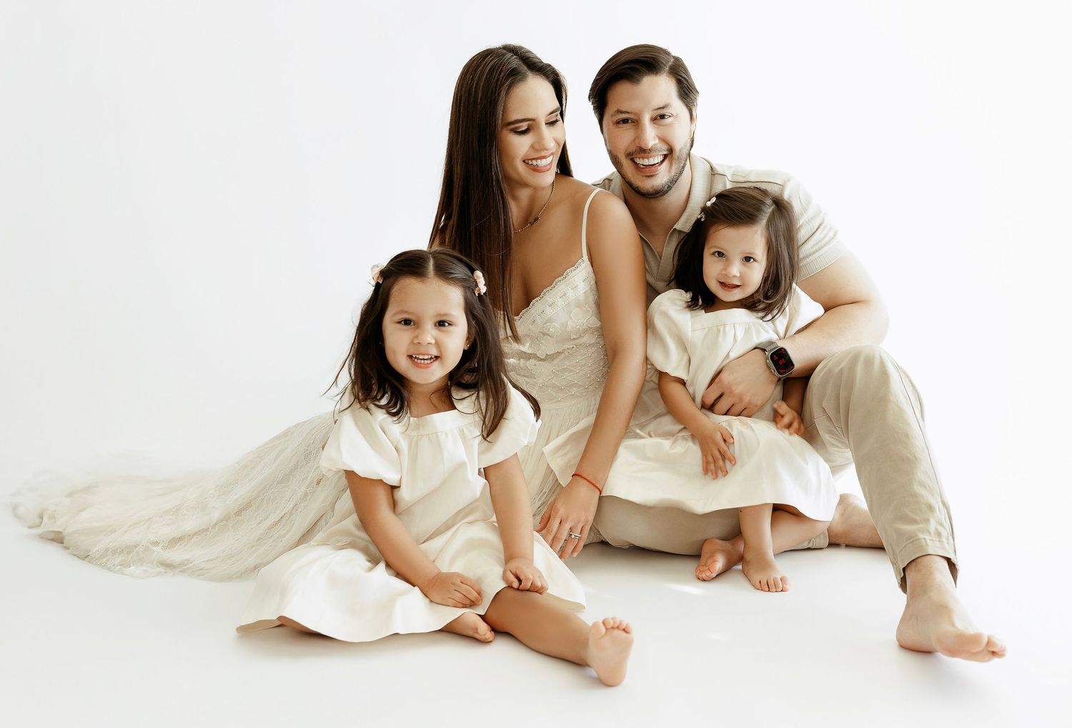 Family wearing white outfits sits together on white backdrop for studio portrait session.