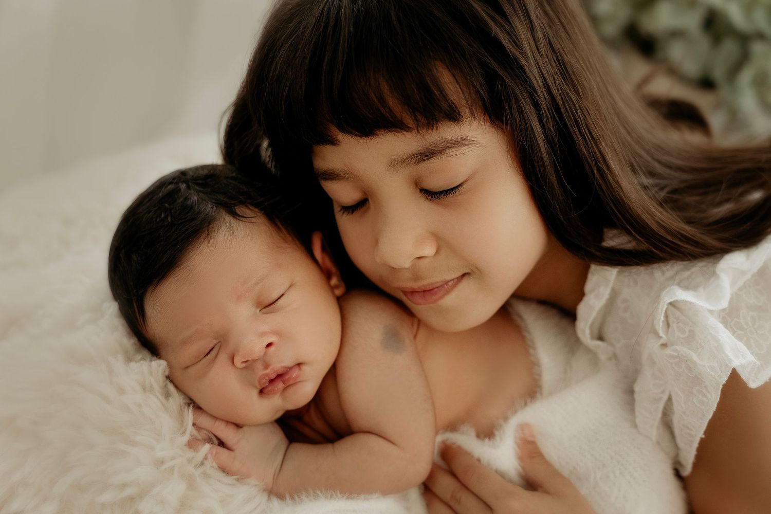A young girl tenderly cradles a sleeping newborn baby on a soft white blanket in a serene studio portrait.