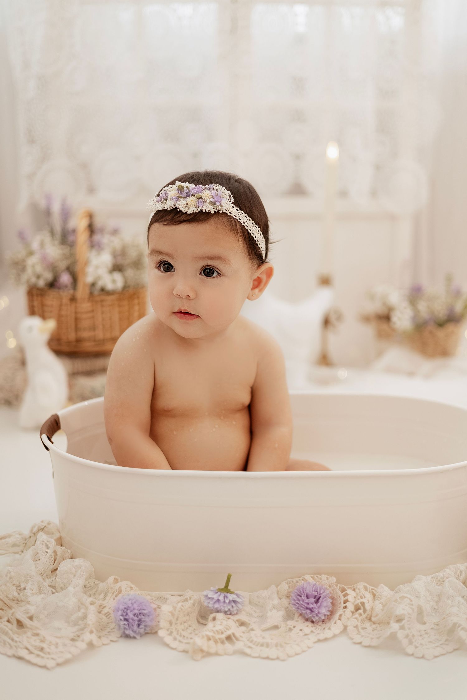 Baby girl in floral headband sits in cream tub, surrounded by lace and purple flowers in soft white studio.