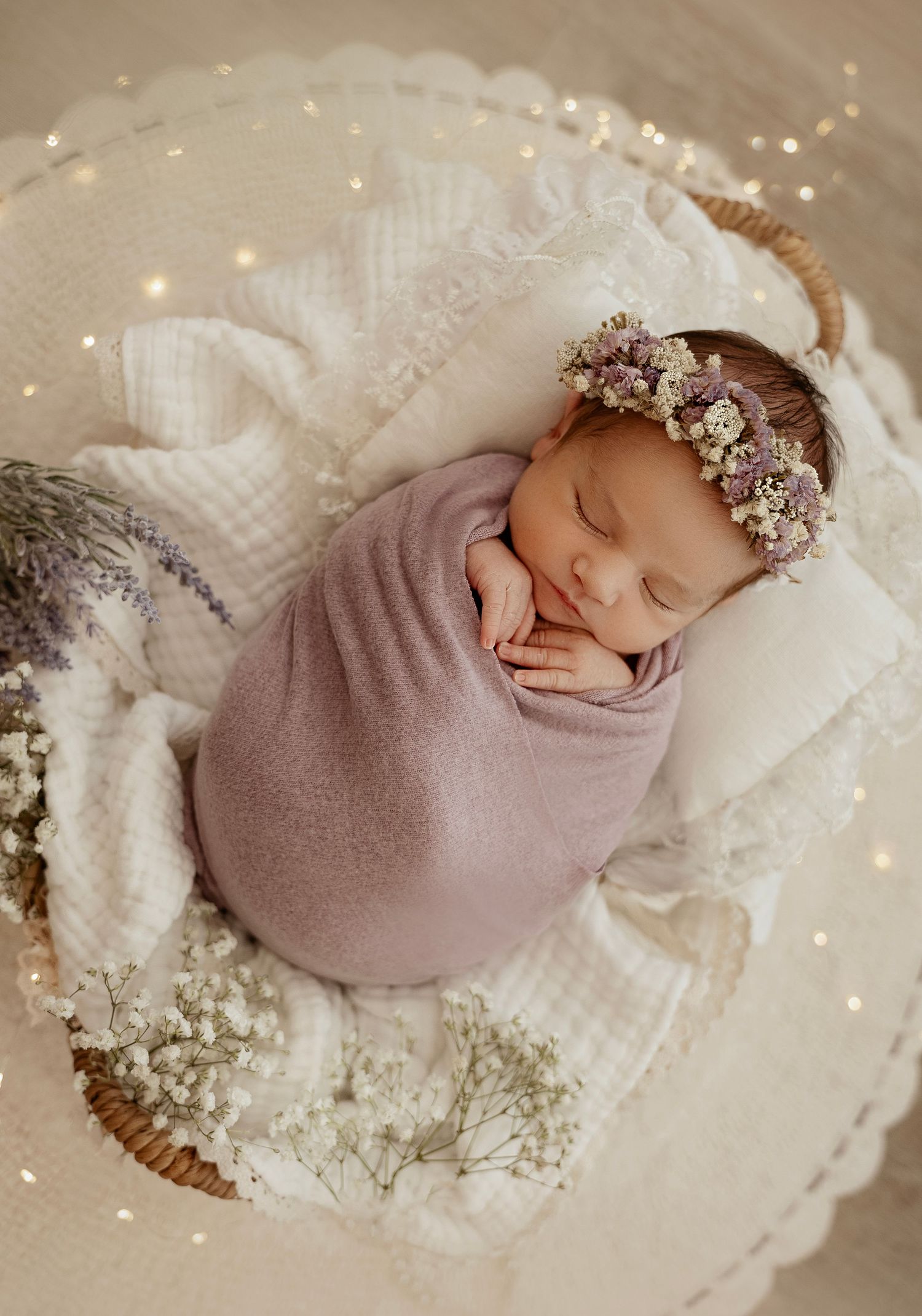 Newborn baby swaddled in mauve wrap on white blanket with delicate floral accents and twinkling lights.
