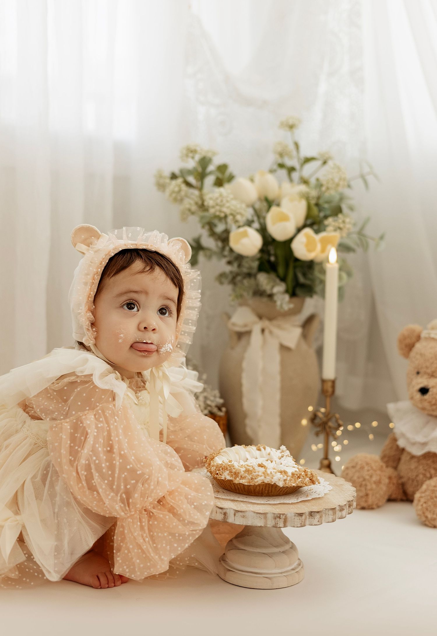 A baby in a delicate peach lace dress poses near white flowers and candles in a dreamy studio setting.