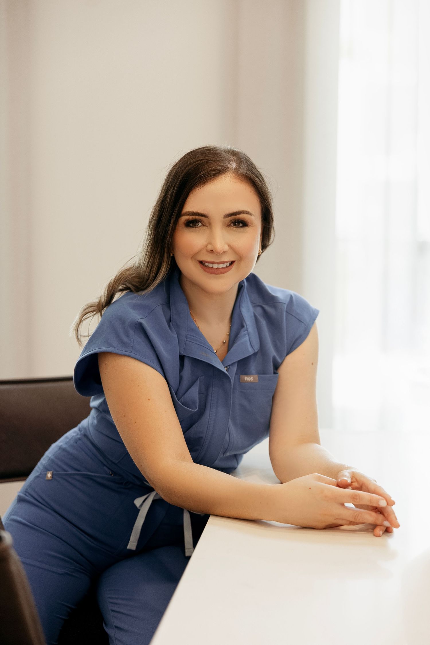Smiling female healthcare professional in blue scrubs leaning on white desk in bright medical office.