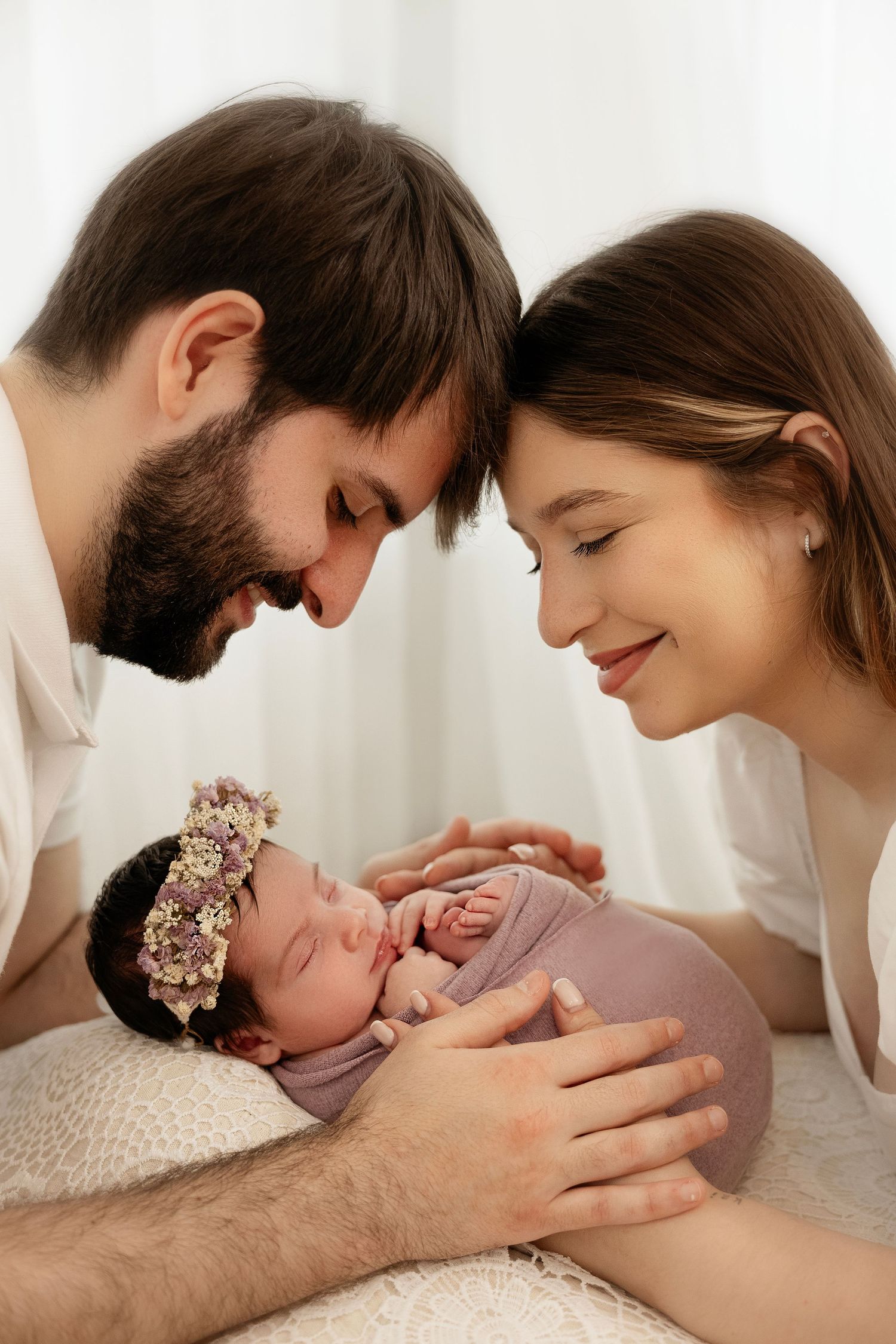 Parents lovingly gaze at their sleeping newborn baby wearing a floral crown in tender family moment.