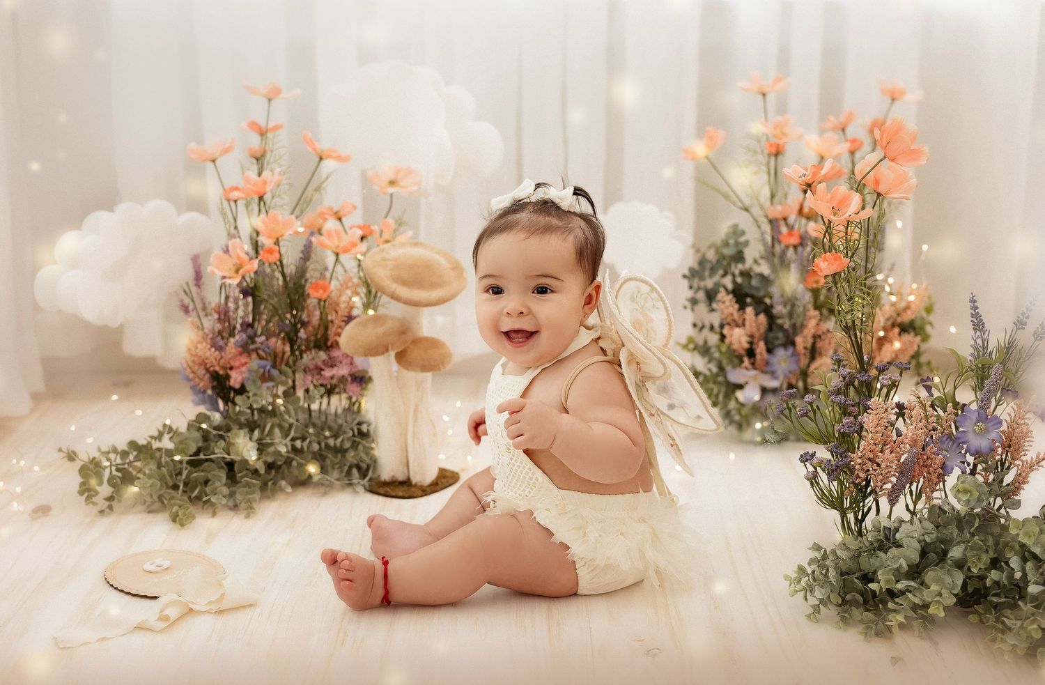 Smiling baby girl in white romper sitting among colorful flowers and mushrooms in a whimsical fairy garden photo setup.