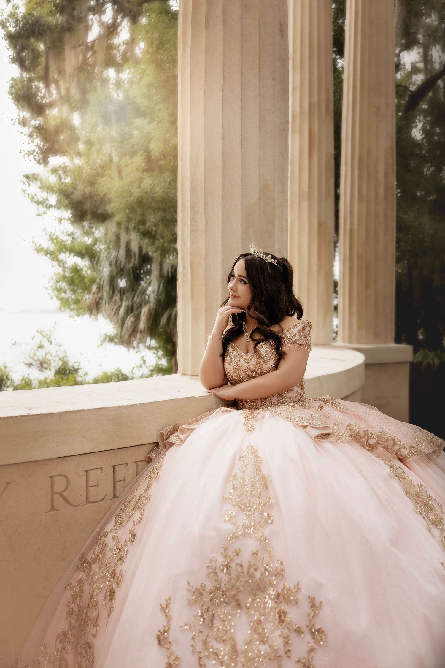 A quinceañera dress in blush pink with ornate gold embellishments is showcased in an elegant columned setting.