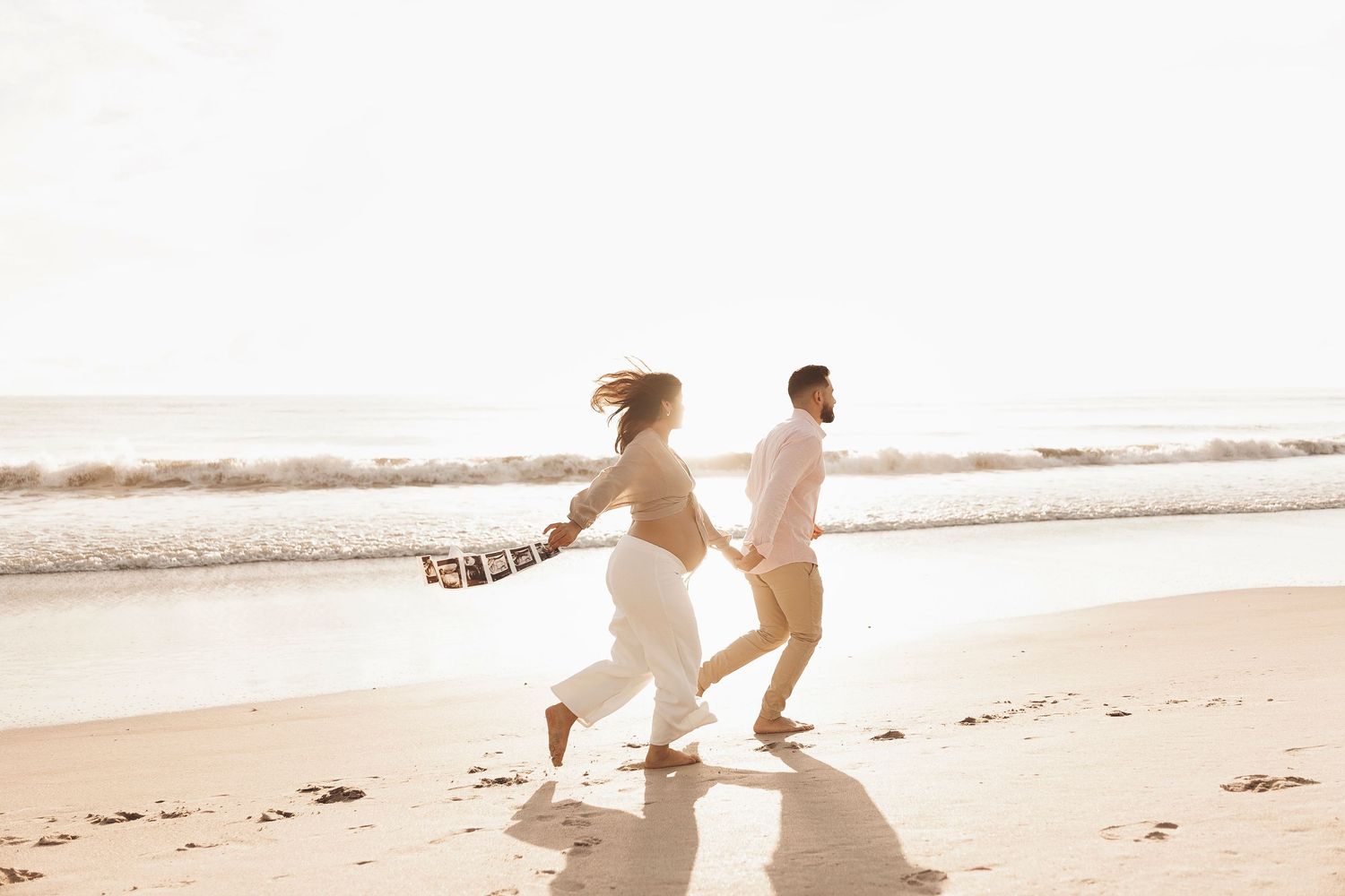 A couple runs playfully along a sandy beach at sunset holding hands while wearing light colored clothing.