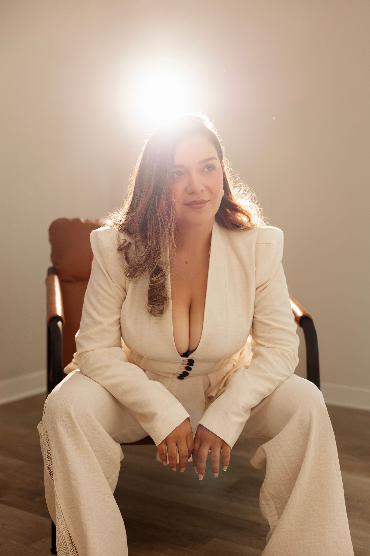 Woman in white blazer suit seated on brown chair with bright backlight in minimalist studio setting.