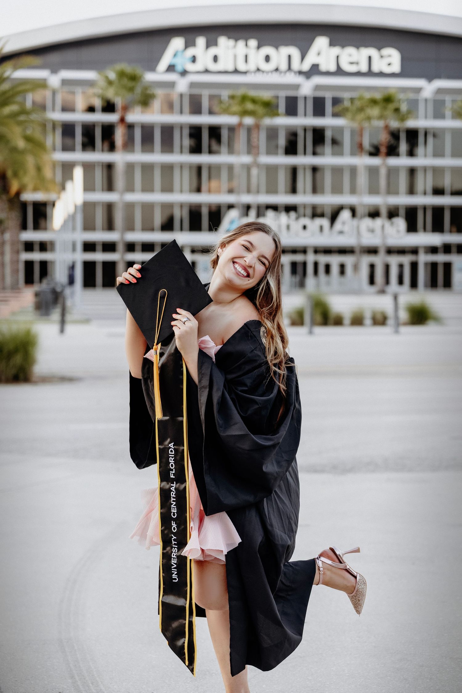 Smiling graduate in black cap and gown poses outside Addition Arena holding her University of Central Florida stole.