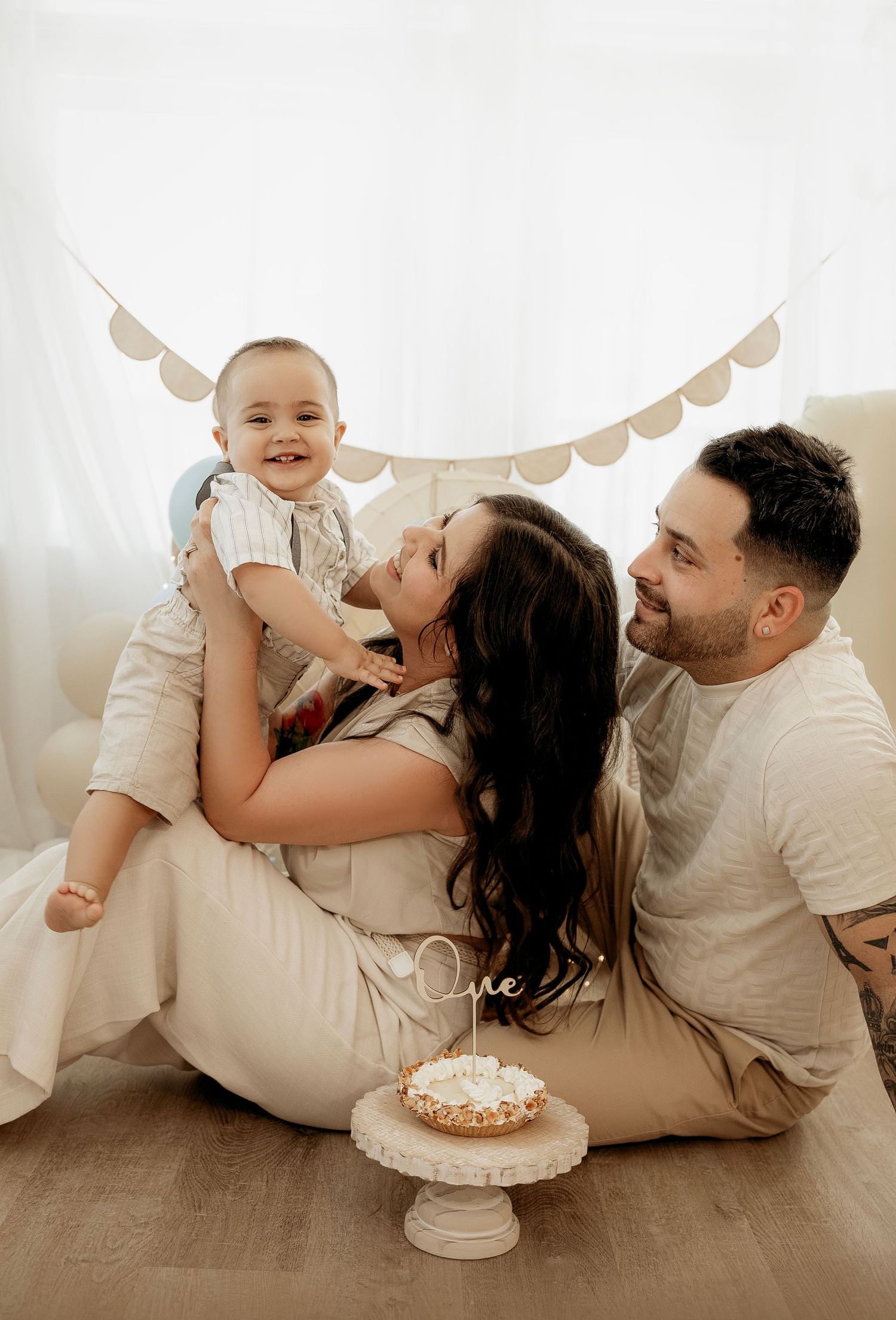 A family wearing cream colored clothes plays together on the floor while posing with a cake for a photo shoot.