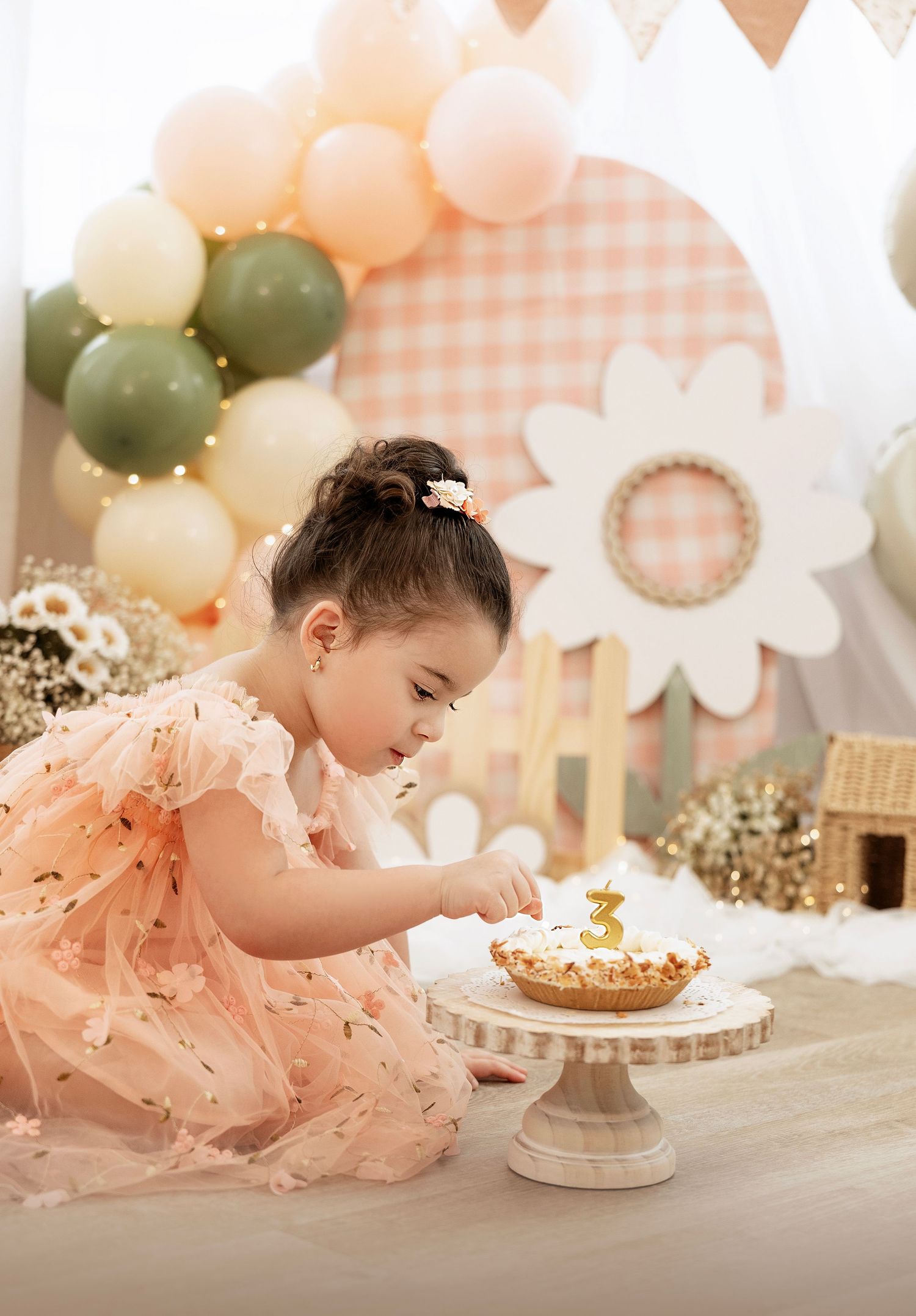 A child in a peach tulle dress reaches for a small cake on a table decorated with pink and white party decor.