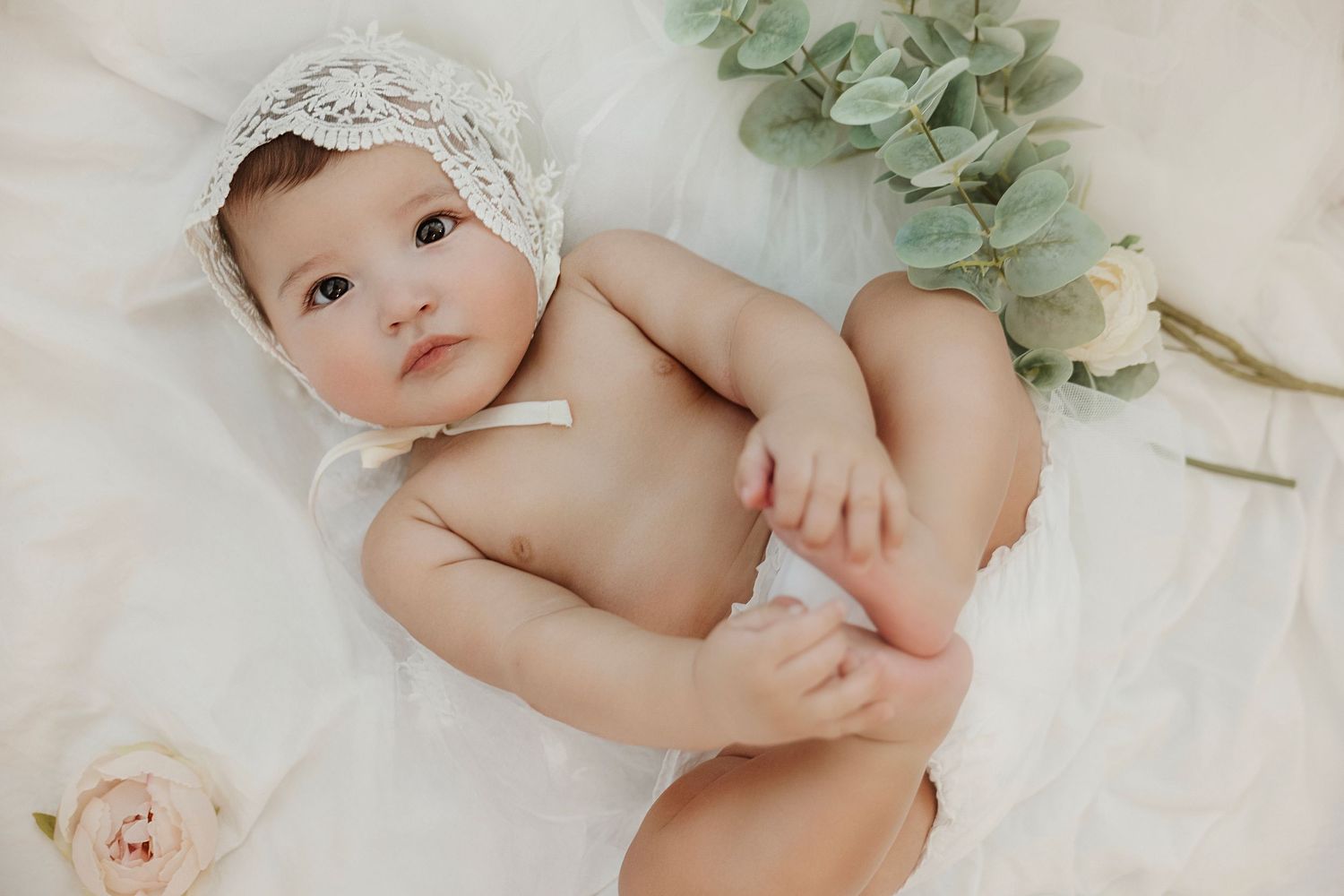 Adorable baby girl wearing a white lace bonnet lying on a soft white blanket with pink roses and eucalyptus.