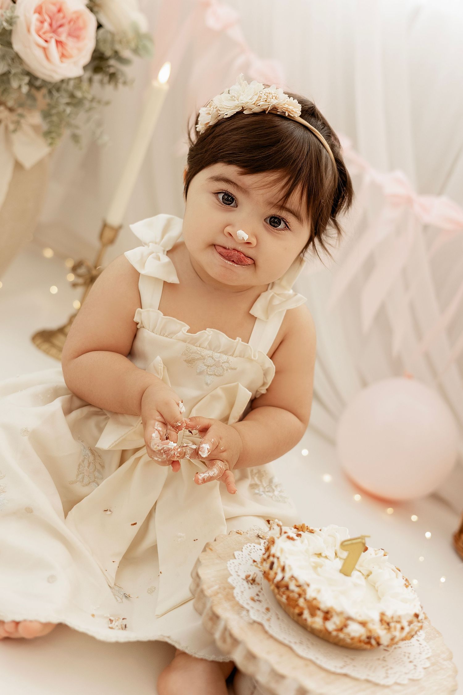 Elegant first birthday celebration setup with white dress, tiara, and decorated cake in soft pink and white tones.