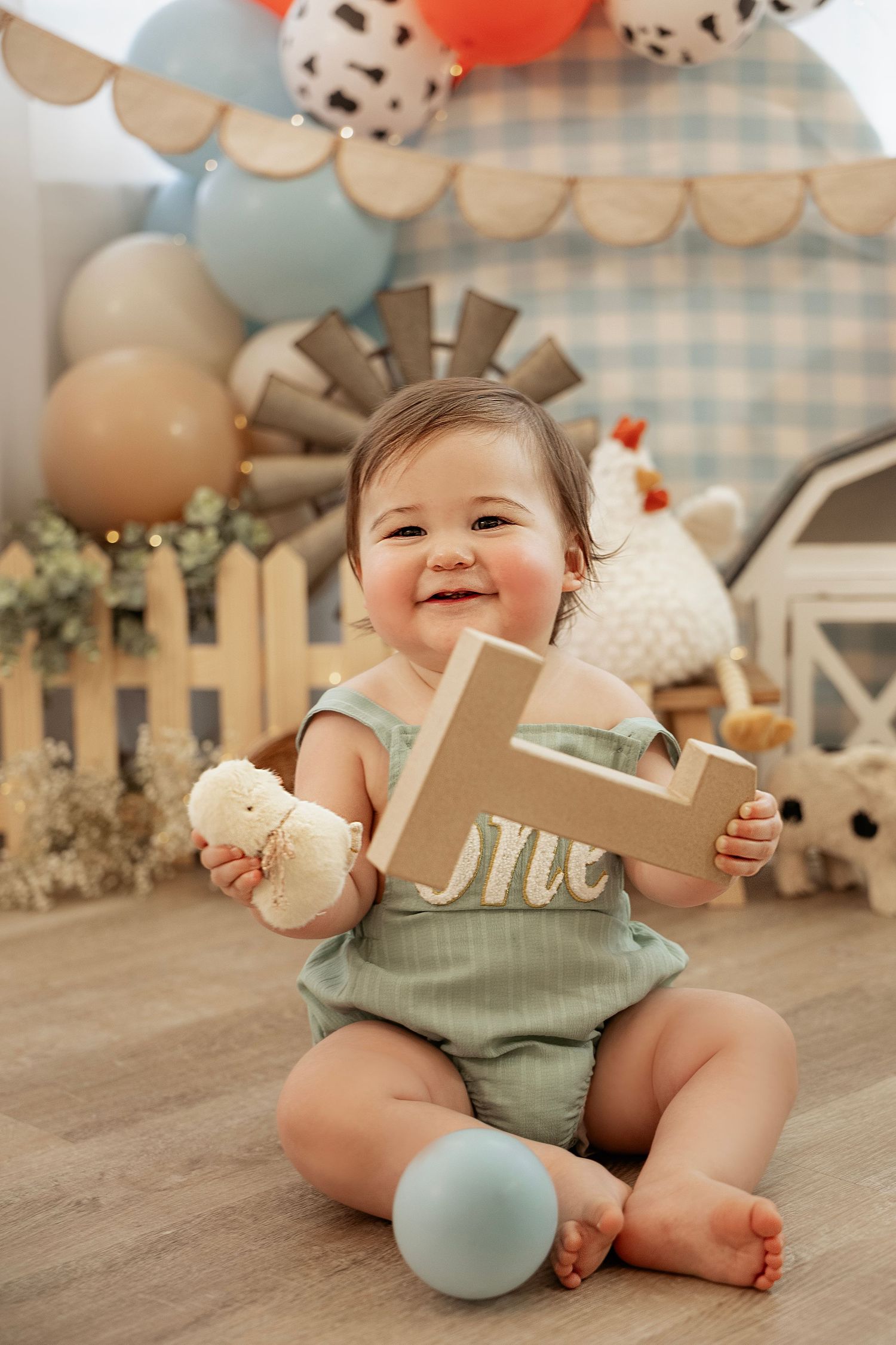 Baby in mint green romper sitting on wooden floor with birthday decorations and balloons in soft pastel colors.