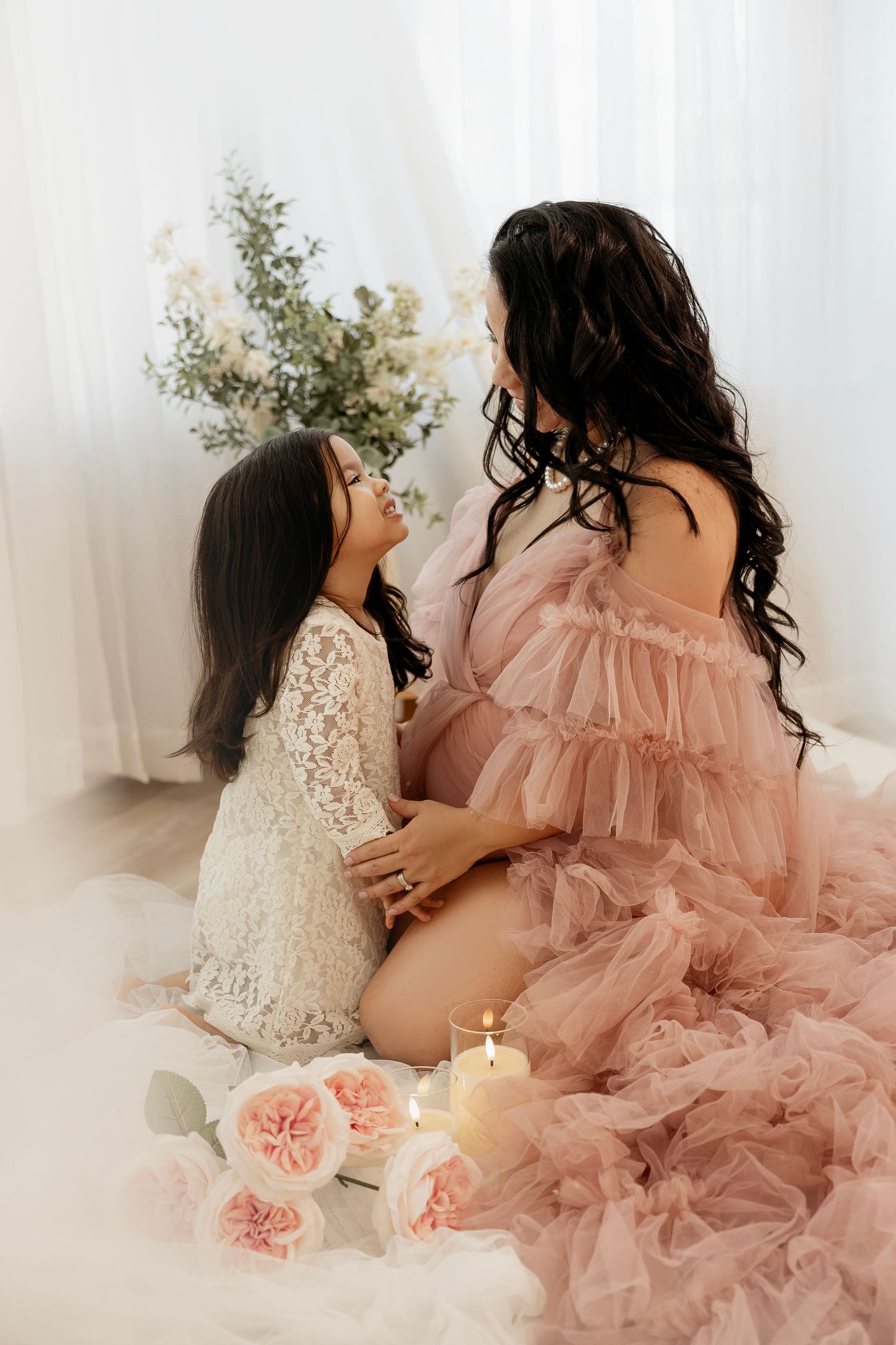 A dreamy maternity photoshoot featuring flowing pink tulle dress and white lace gown against soft white curtains.