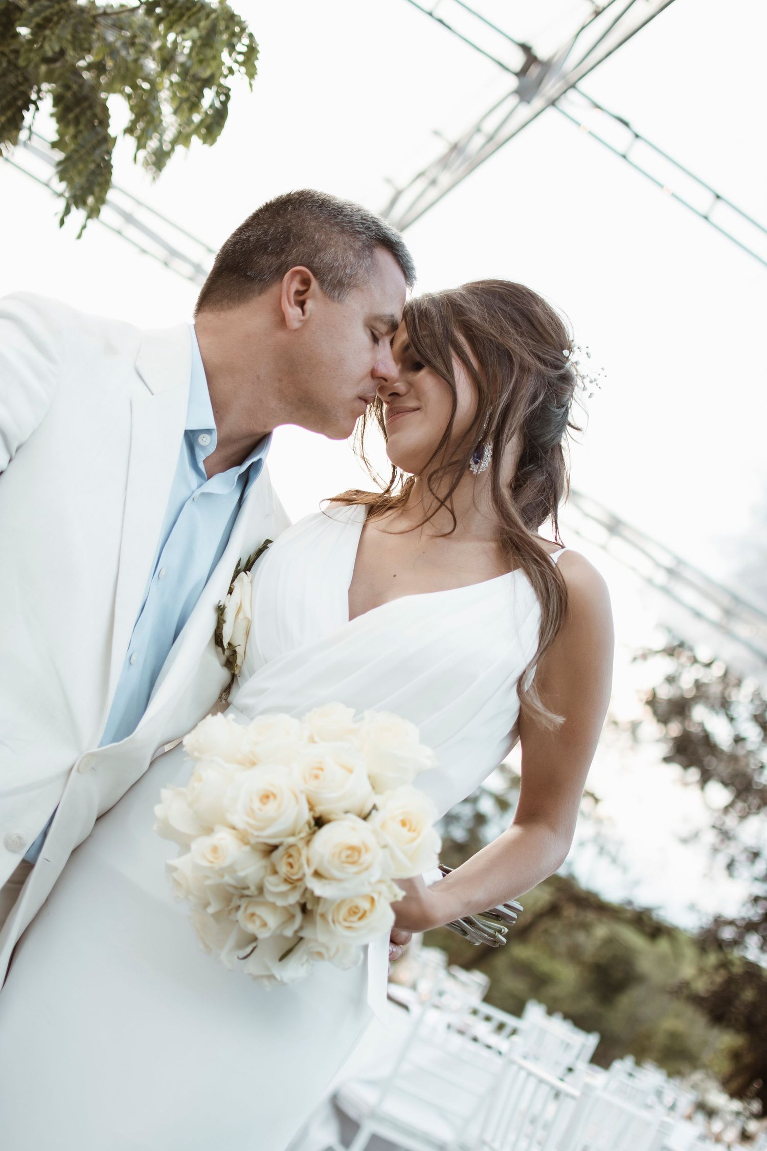 A romantic wedding couple shares intimate moments in a bright, airy setting with white roses and elegant attire.