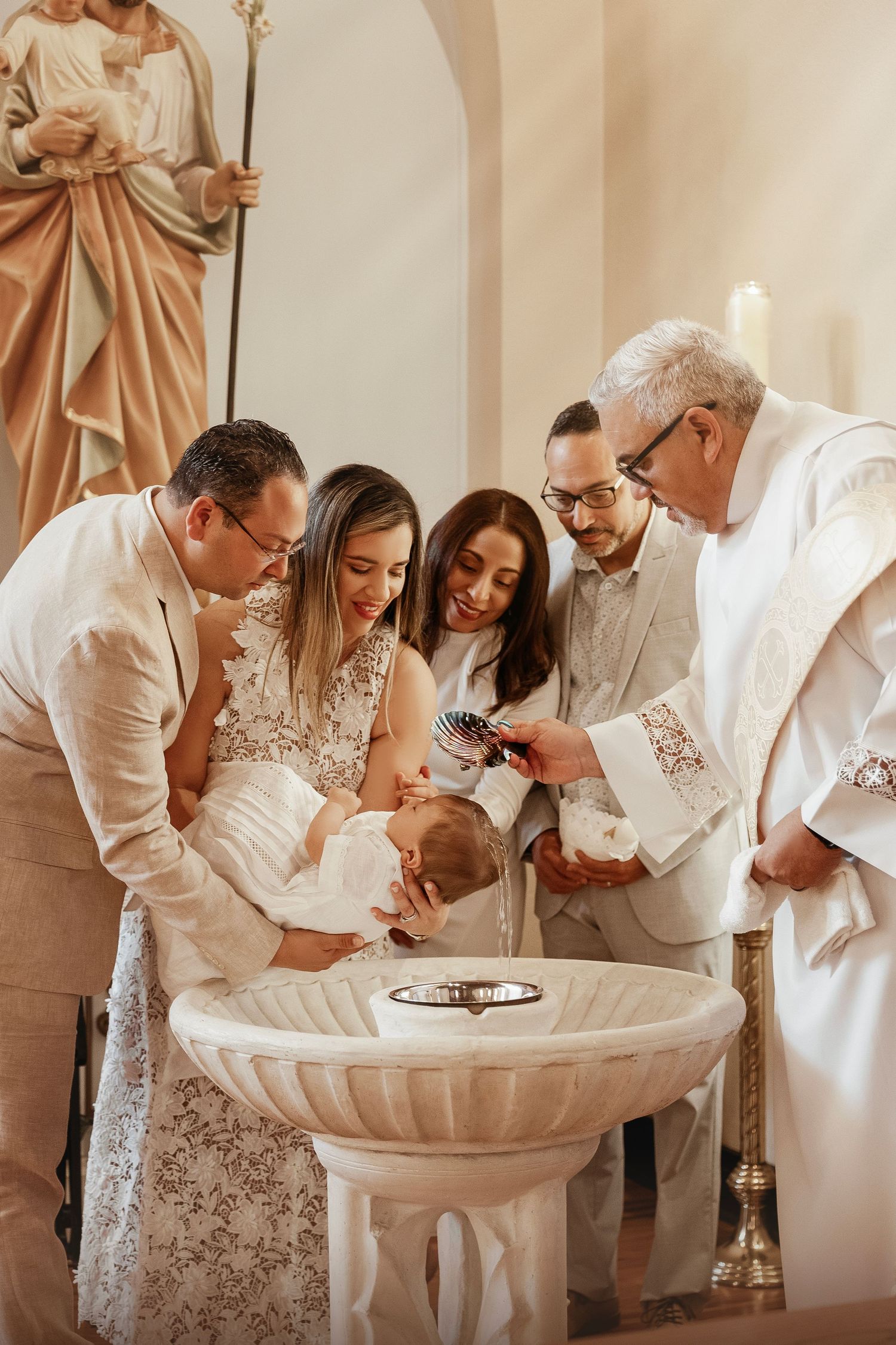 A baptism ceremony takes place at a church font as family members and priest gather around during the sacred moment.