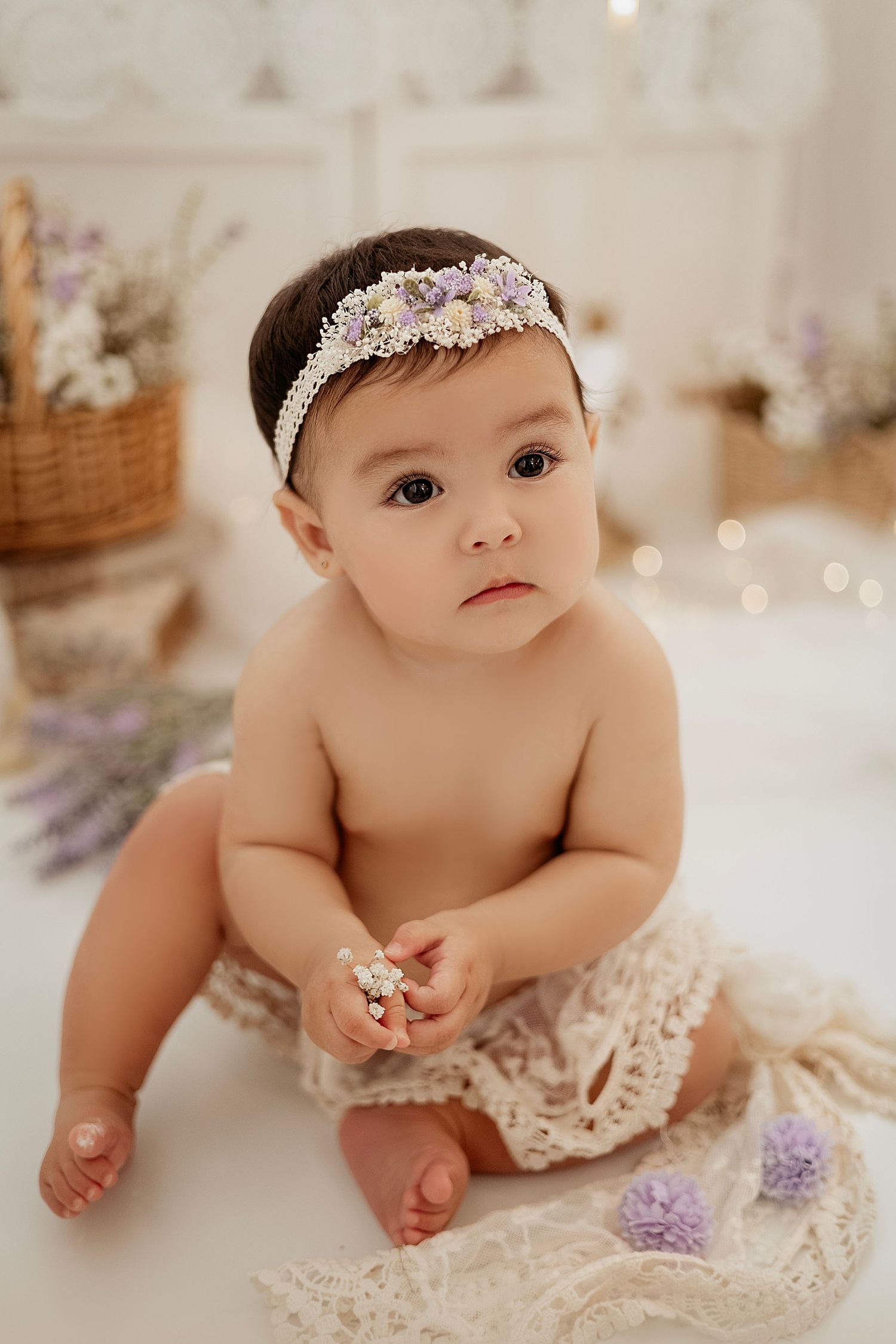 Baby girl in lace bloomers and floral headband sits on white backdrop with lavender flowers and wicker basket.