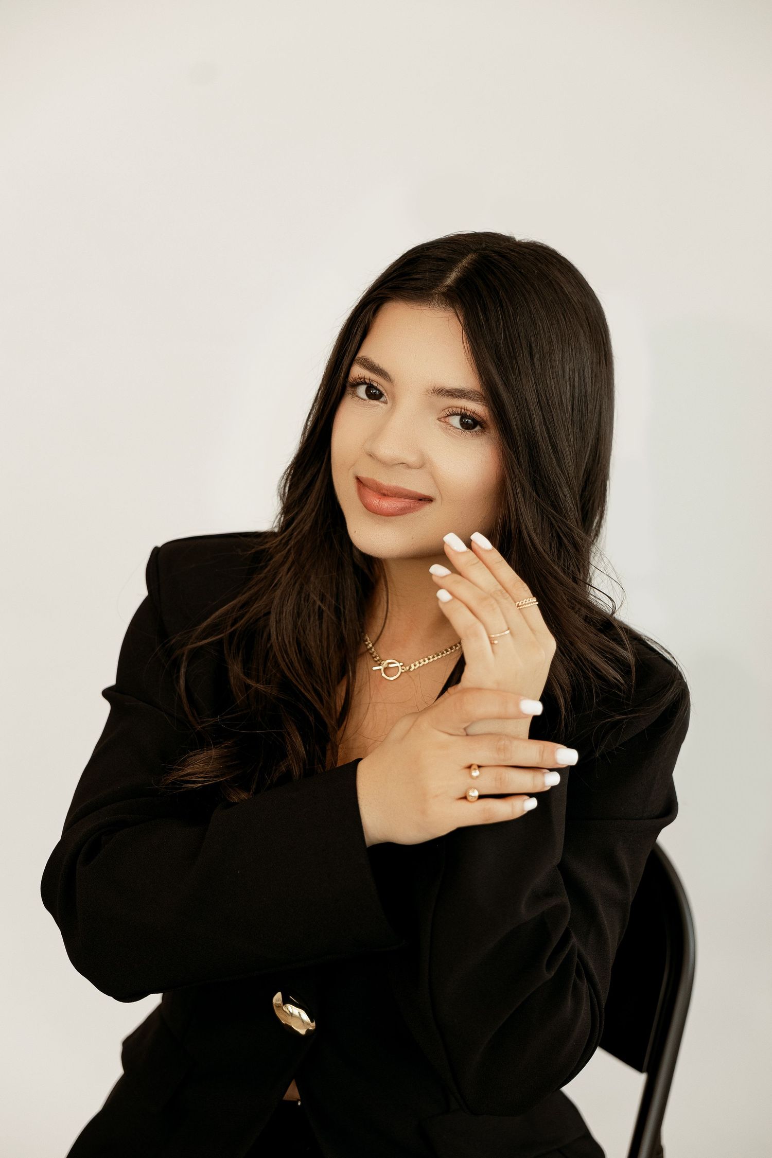 Professional studio portrait of someone in a black blazer with long dark hair and jewelry against a light background.