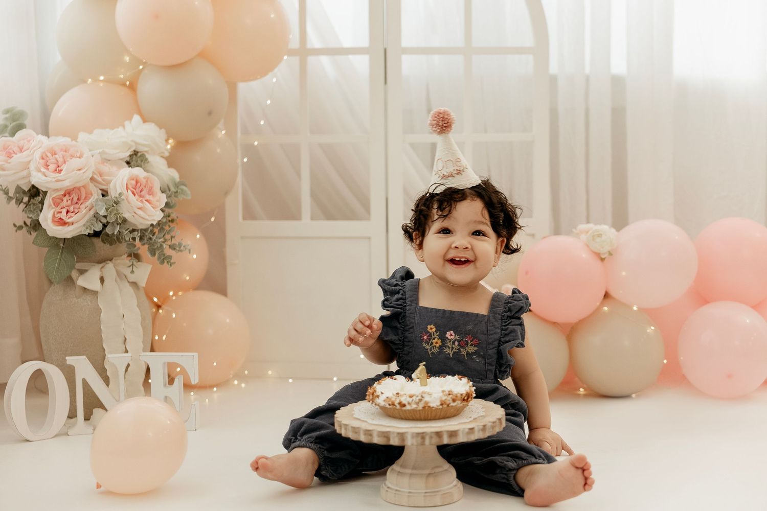 First birthday celebration setup with pink balloons, floral arrangements and cake smash scene in soft pastel colors.