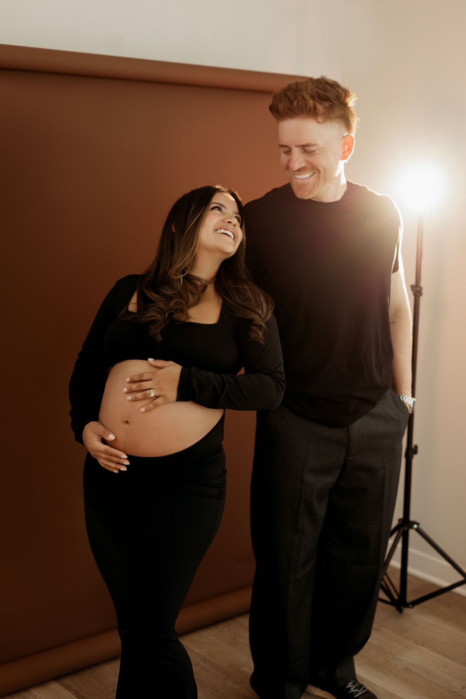A couple shares intimate moments during a maternity photoshoot in warm lighting against a brown backdrop.