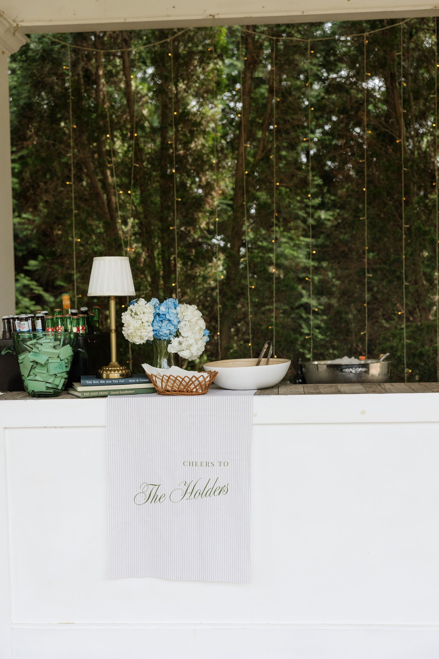 Elegant outdoor wedding bar setup with hydrangeas, brass lamp, and personalized The Holders runner amid string lights.