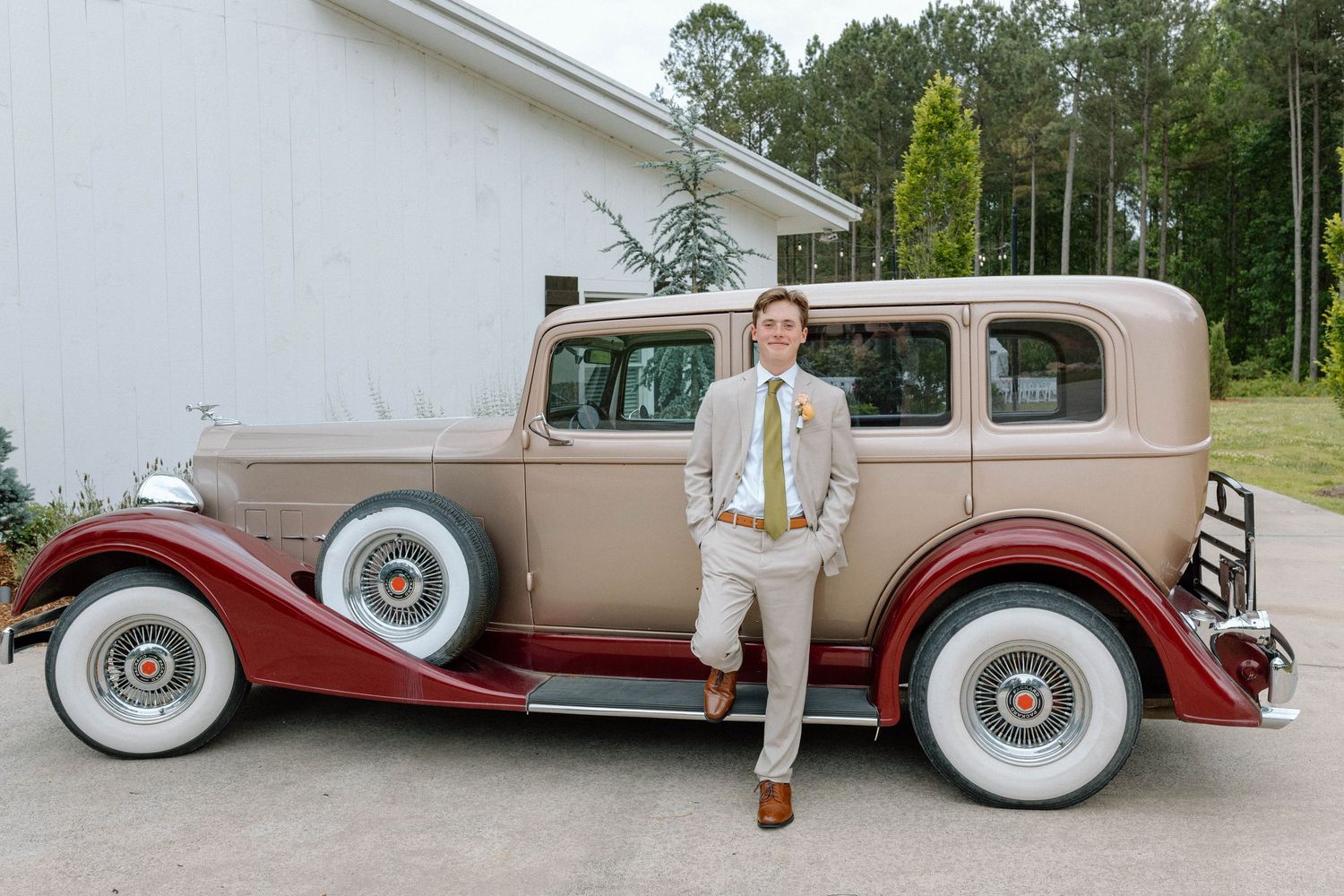 Groom in tan suit poses beside a vintage brown and red classic car outside a white building.