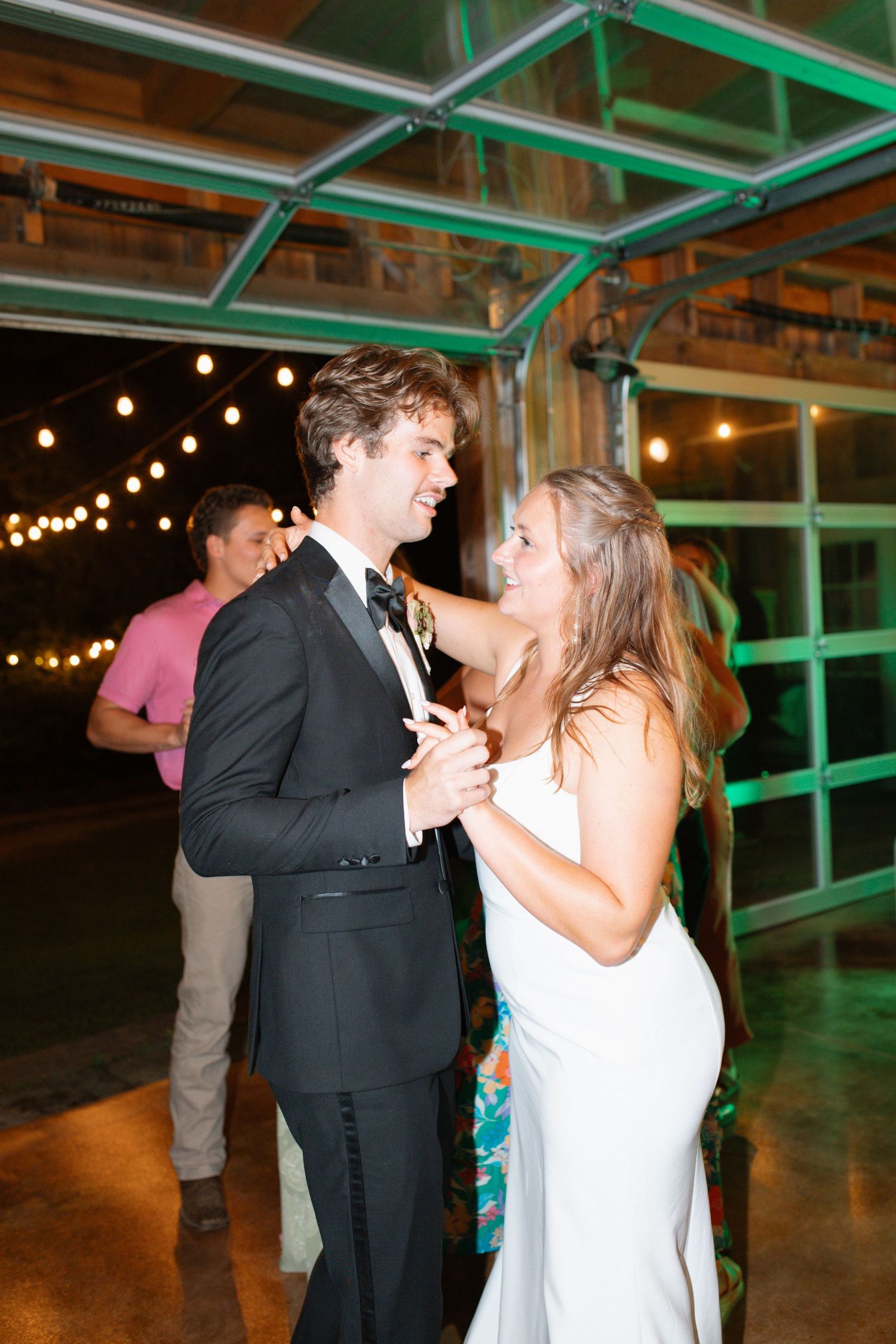 Bride in white gown and groom in black tuxedo share romantic first dance under string lights at reception.